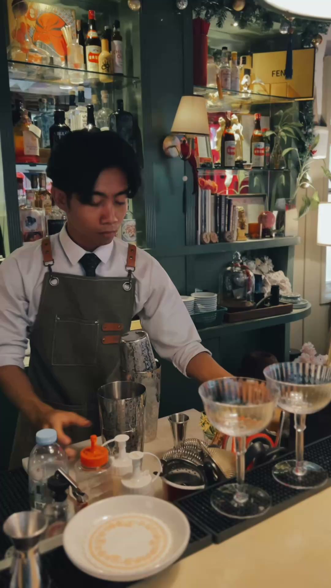 บาร์เทนเดอร์สวมผ้ากันเปื้อนกำลังทำค็อกเทลหลังเคาน์เตอร์บาร์พร้อมอุปกรณ์ผสมเครื่องดื่มและแก้วค็อกเทล