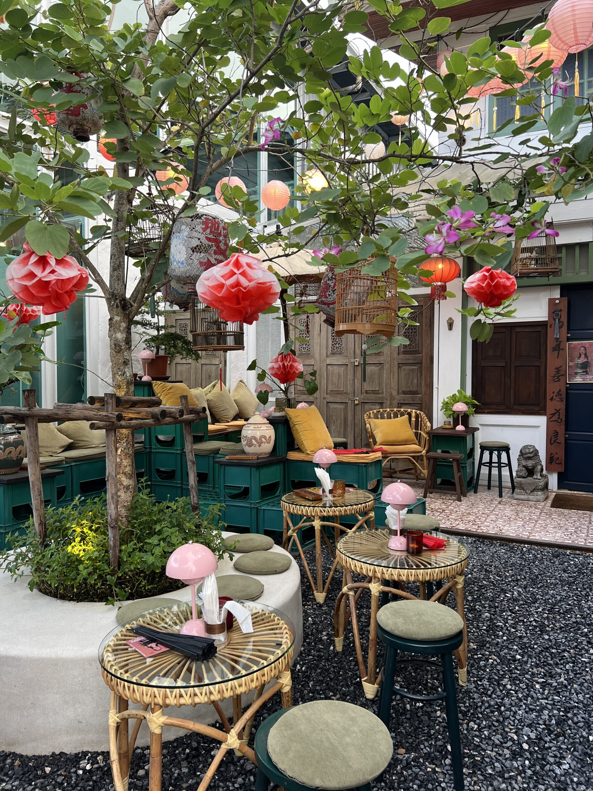 ลานคาเฟ่สไตล์จีนตกแต่งด้วยโคมไฟสีแดงและชมพู โต๊ะหวายกระจก เก้าอี้หวาย หมอนสีเหลือง ม้านั่งจากลังพลาสติกสีเขียว ใต้ร่มเงาต้นไม้ บรรยากาศสวยงามน่านั่ง