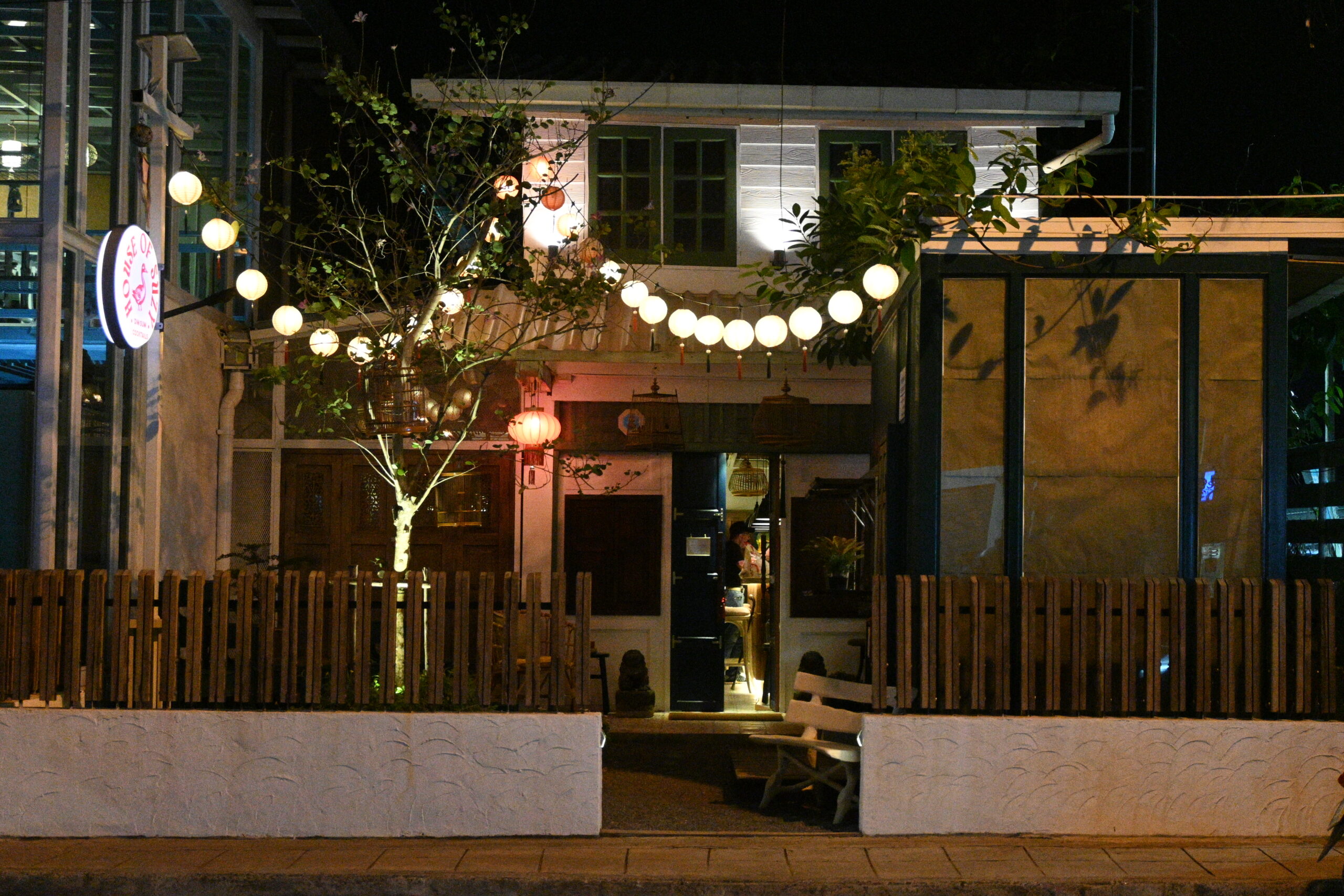 ด้านหน้าร้านคาเฟ่สไตล์วินเทจในเวลากลางคืน ตกแต่งด้วยโคมไฟกลมสีขาวและโคมจีน มีรั้วไม้และต้นไม้ใหญ่หน้าร้าน