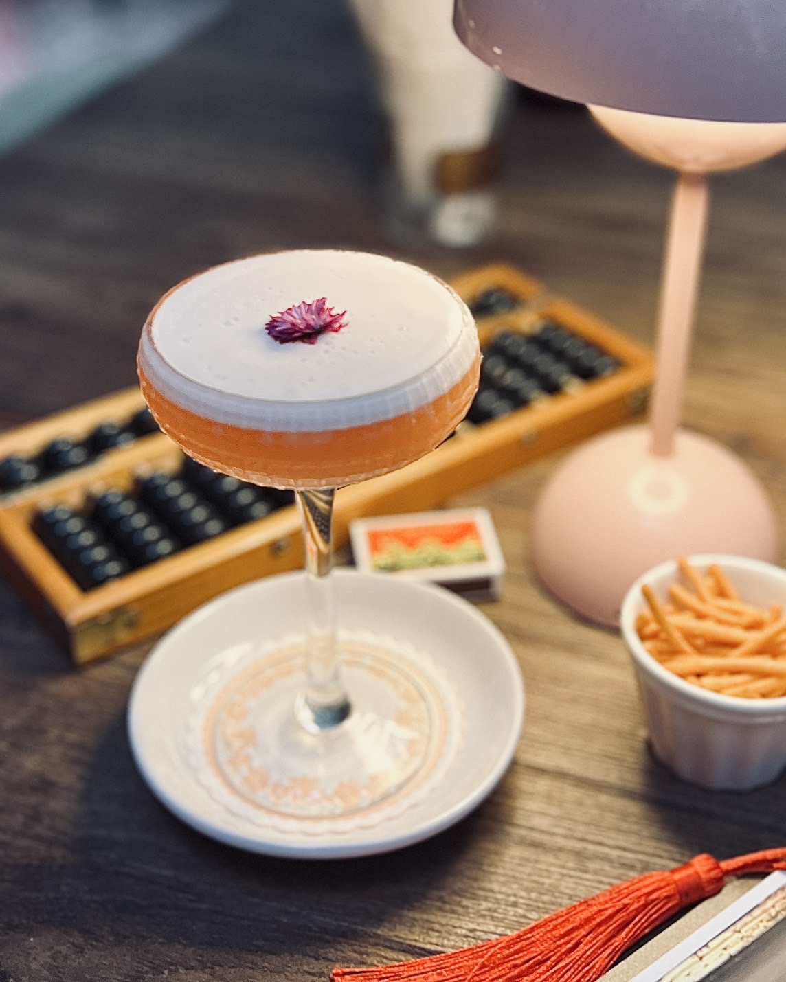 ค็อกเทลสีส้มพร้อมฟองนุ่มด้านบนตกแต่งด้วยดอกไม้สีม่วง เสิร์ฟในแก้วคูเป้บนจานรองสวยงาม พร้อมลูกคิดจีนและโคมไฟสีชมพูเป็นฉากหลัง