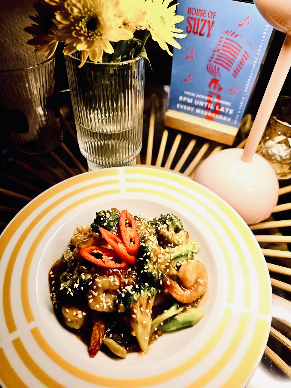 จานผัดผักบร็อคโคลี่กับเม็ดมะม่วงหิมพานต์ราดซอสโปะพริกแดงและงาขาว บนจานลายทางสีเหลืองที่ร้าน House of Suzy พร้อมแจกันดอกไม้และป้ายโปรโมทดนตรีสดทุกวันพุธ