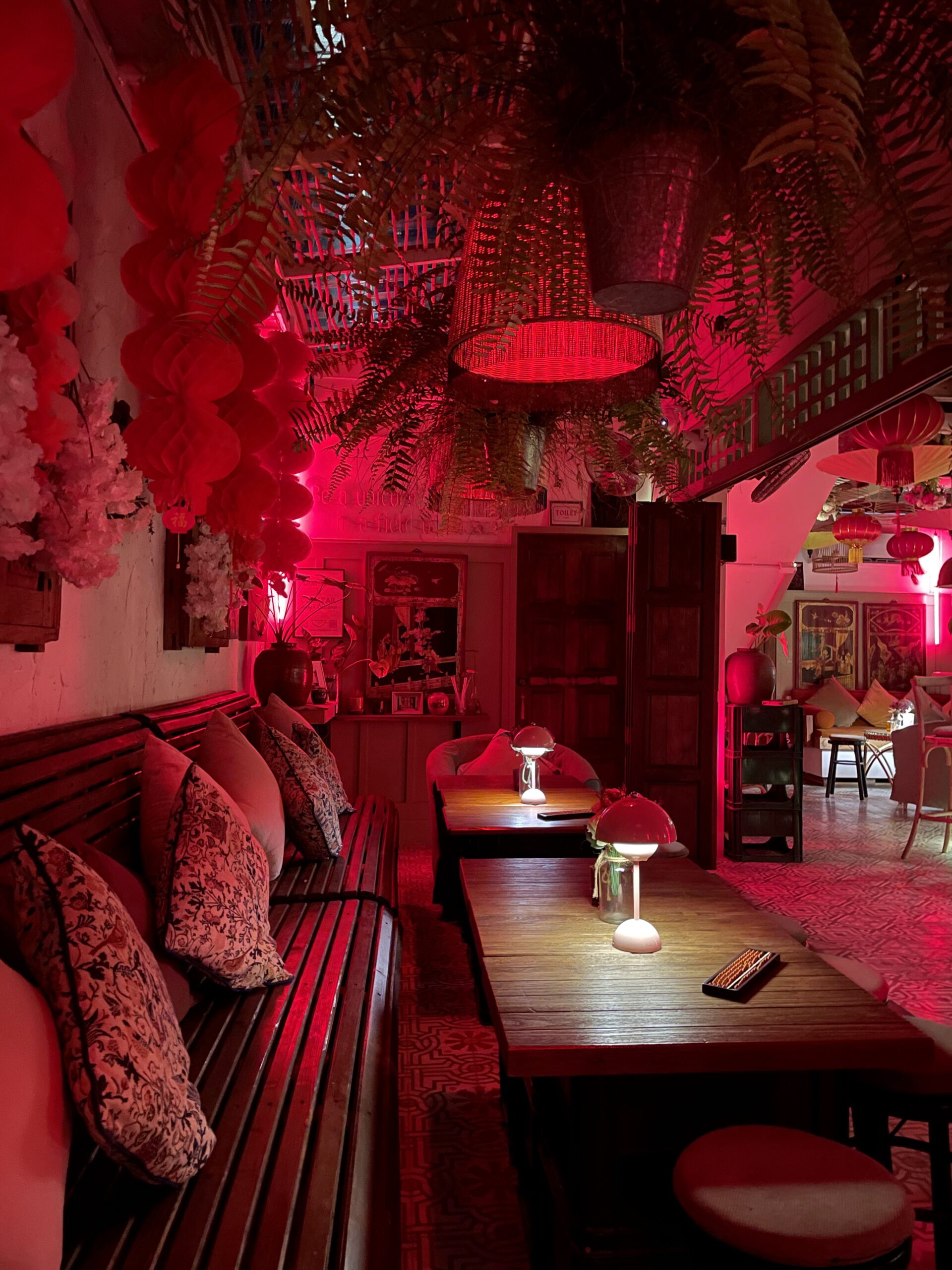 ร้านอาหารบรรยากาศแสงสีแดงตกแต่งสไตล์เอเชียพร้อมโคมไฟหวาย เฟิร์น โคมแดง โซฟาไม้มีหมอนอิง และโต๊ะไม้พร้อมโคมไฟตั้งโต๊ะ