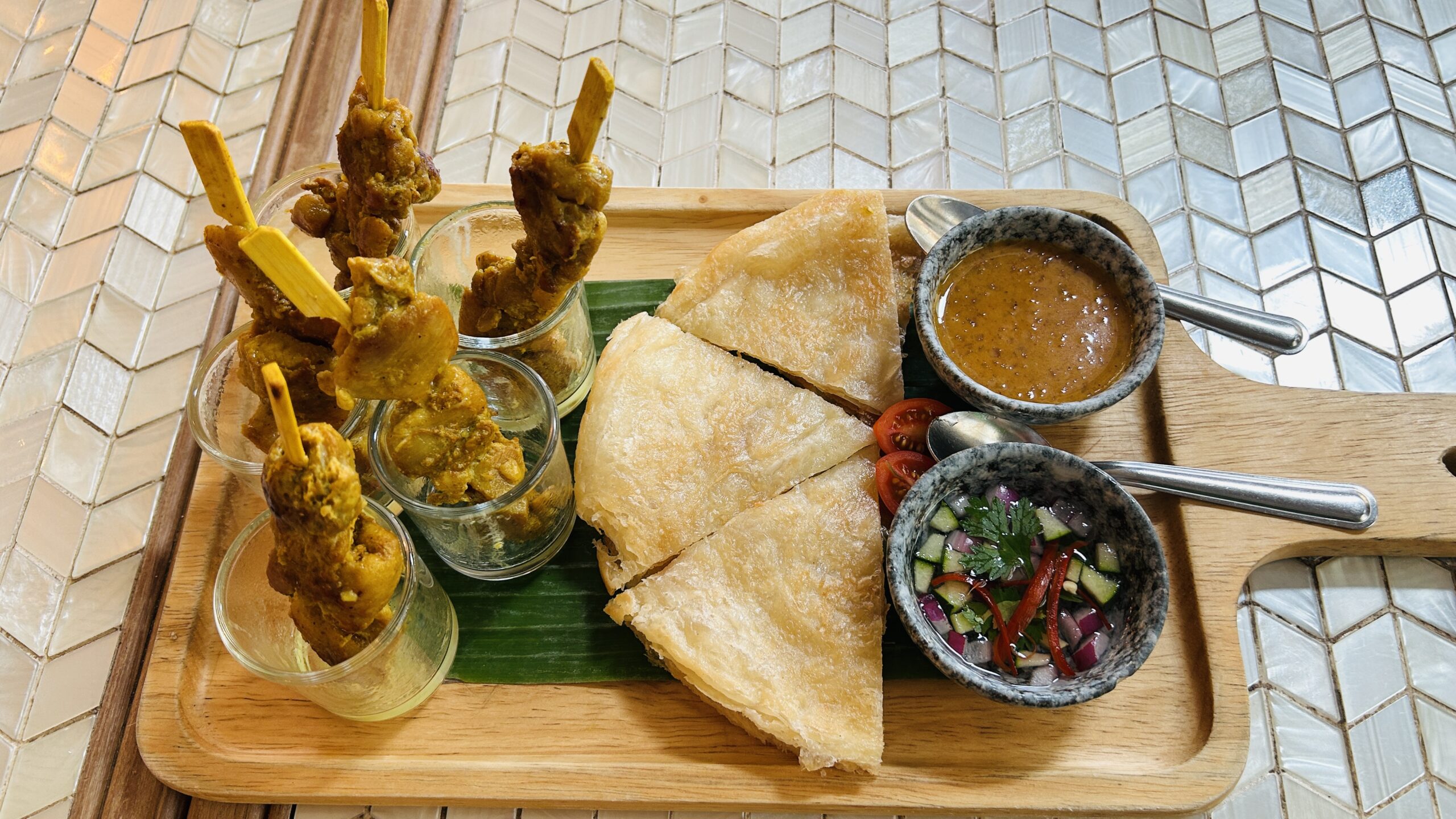 จานไม้เสิร์ฟสะเต๊ะไก่เสียบไม้ในแก้วใส พร้อมโรตีแผ่นกรอบ น้ำจิ้มสะเต๊ะ และอาจาดแตงกวาหอมแดง