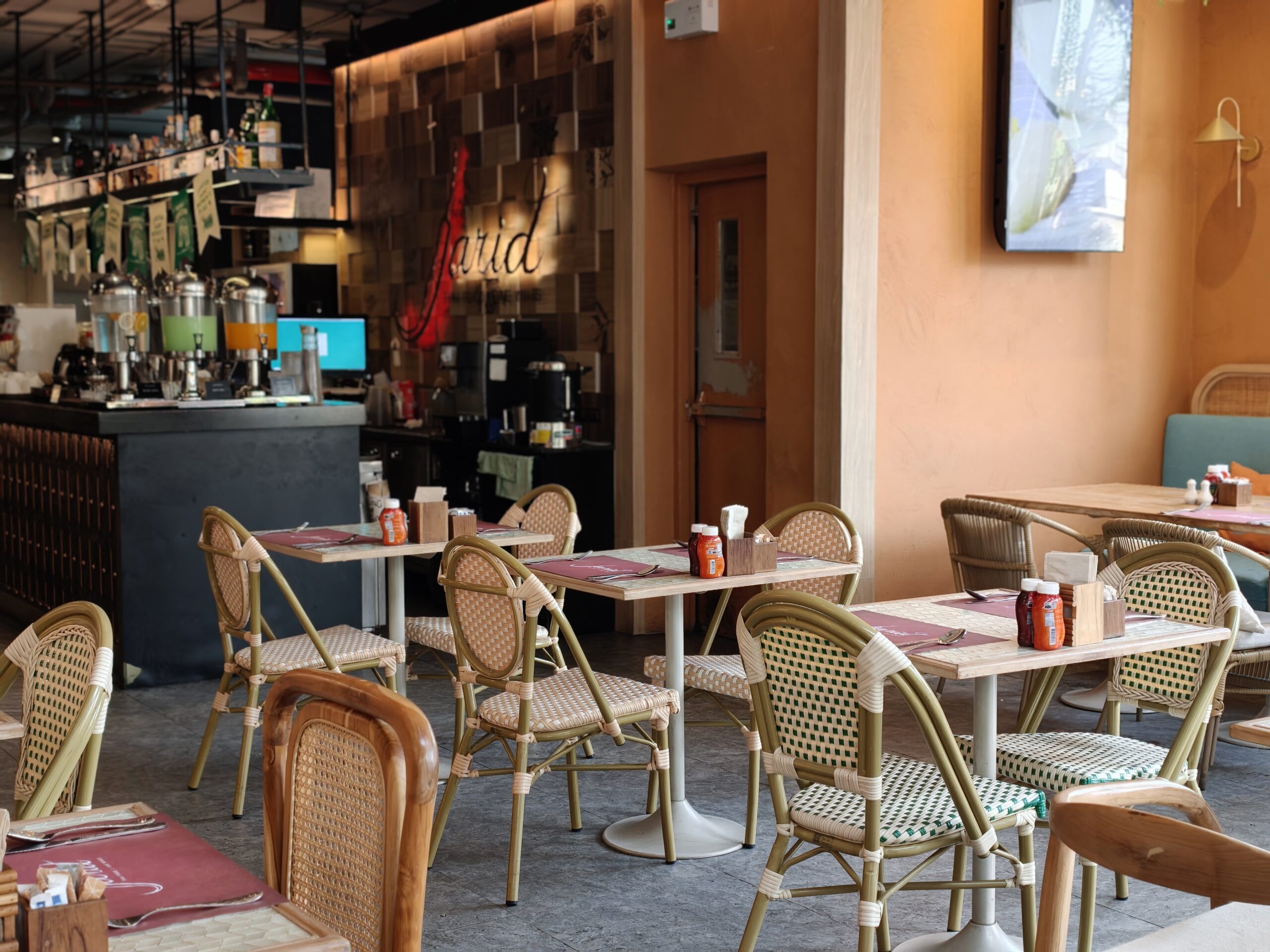 บรรยากาศภายในร้านอาหารสไตล์บิสโทรที่มีโต๊ะเก้าอี้หวายจัดเรียงอย่างน่านั่ง พร้อมเคาน์เตอร์บาร์และเครื่องดื่มด้านหลัง