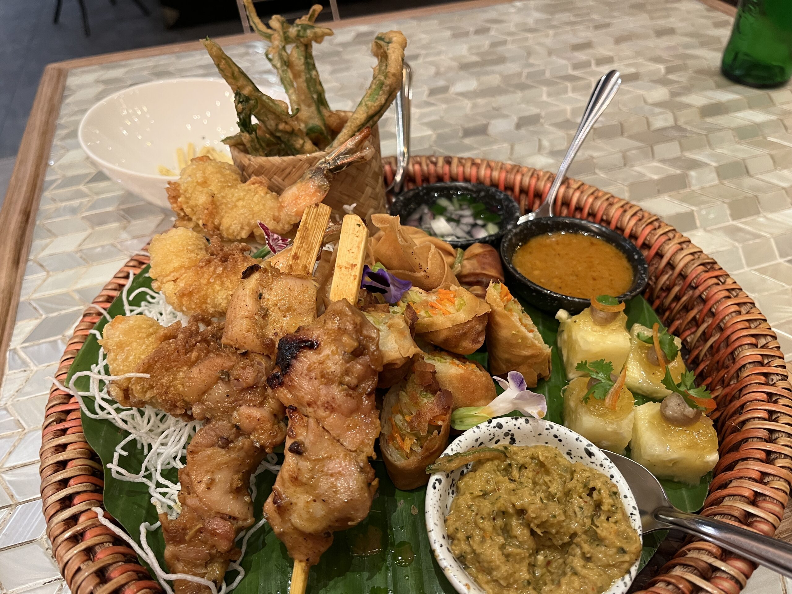 จานรวมอาหารเรียกน้ำย่อยไทยในตะกร้าหวาย มีไก่สะเต๊ะ กุ้งทอด ปอเปี๊ยะทอด ผักชุบแป้งทอด พร้อมน้ำจิ้มหลายชนิด บนใบตอง