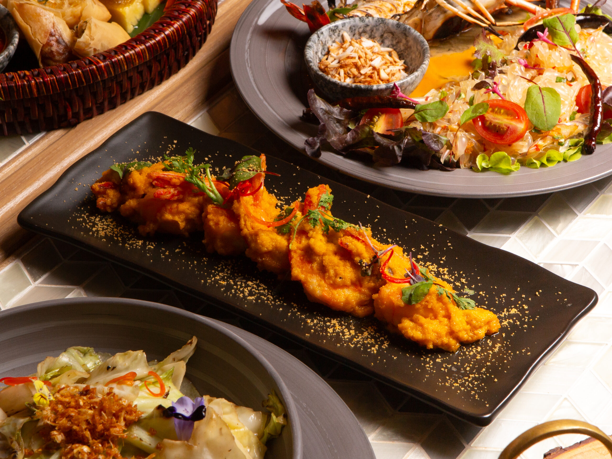 จานอาหารไทยหลากหลายบนโต๊ะ มีกุ้งทอดซอสไข่เค็มบนจานดำ สลัดกุ้งแม่น้ำ ผักผัดน้ำมันหอย และขนมจีบในตะกร้า จัดอย่างสวยงาม