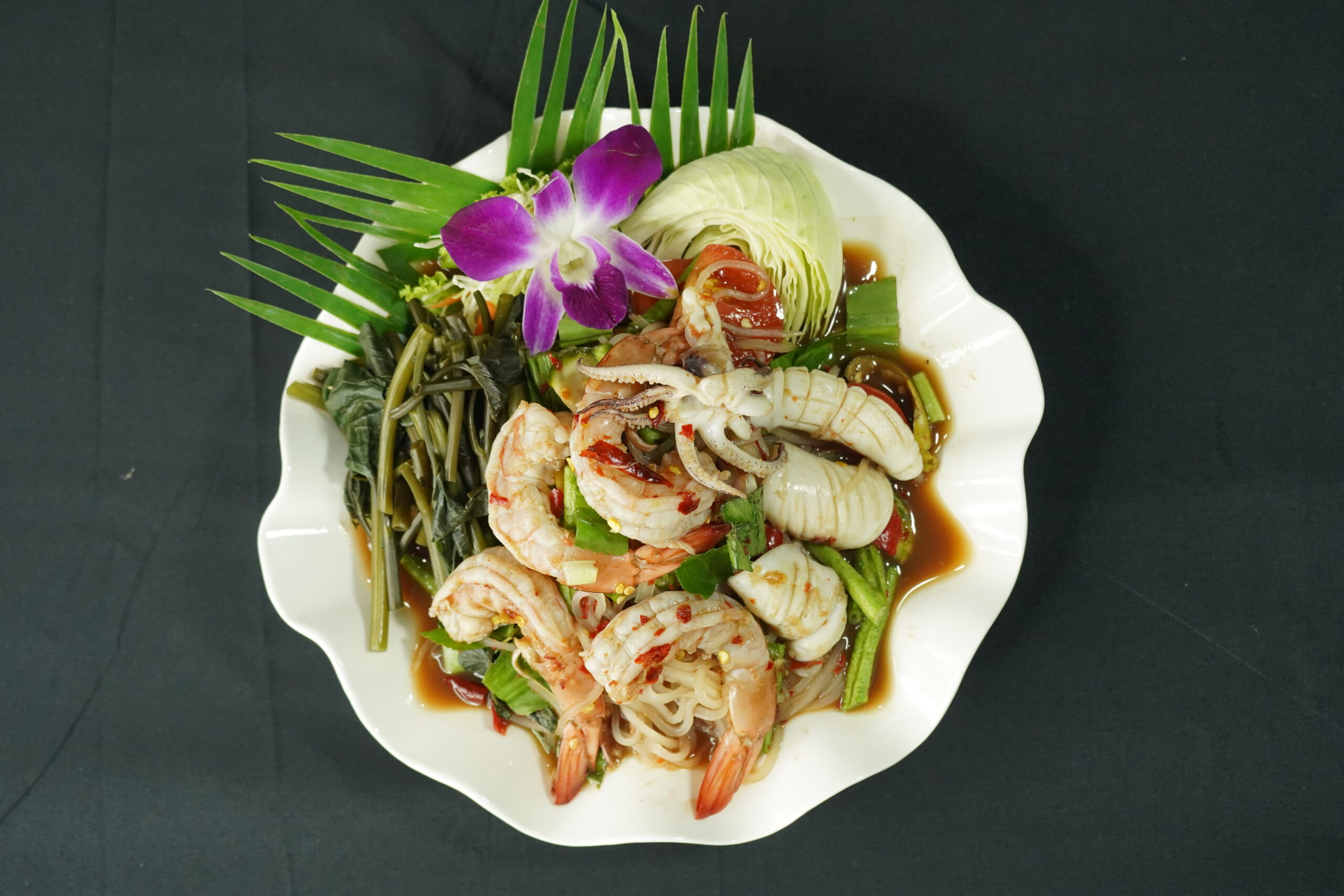 ยำทะเลรวมมิตรกุ้ง ปลาหมึก ราดน้ำยำรสจัด จัดจานสวยด้วยดอกกล้วยไม้ม่วงและผักสด บนจานขาวรูปเปลือกหอย
