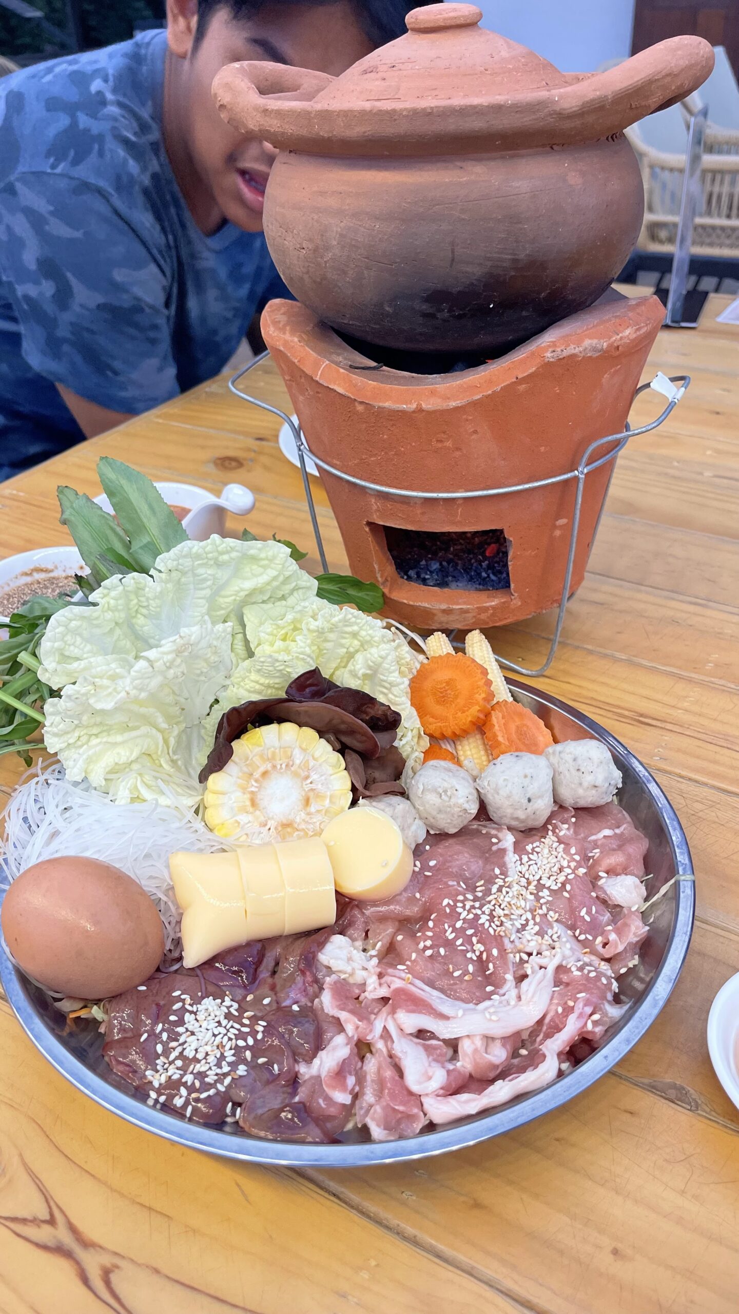 ชุดจิ้มจุ่มหม้อดินพร้อมเนื้อหมูสไลซ์โรยงา ลูกชิ้น ผัก ข้าวโพด วุ้นเส้น เต้าหู้ และไข่ บนโต๊ะไม้