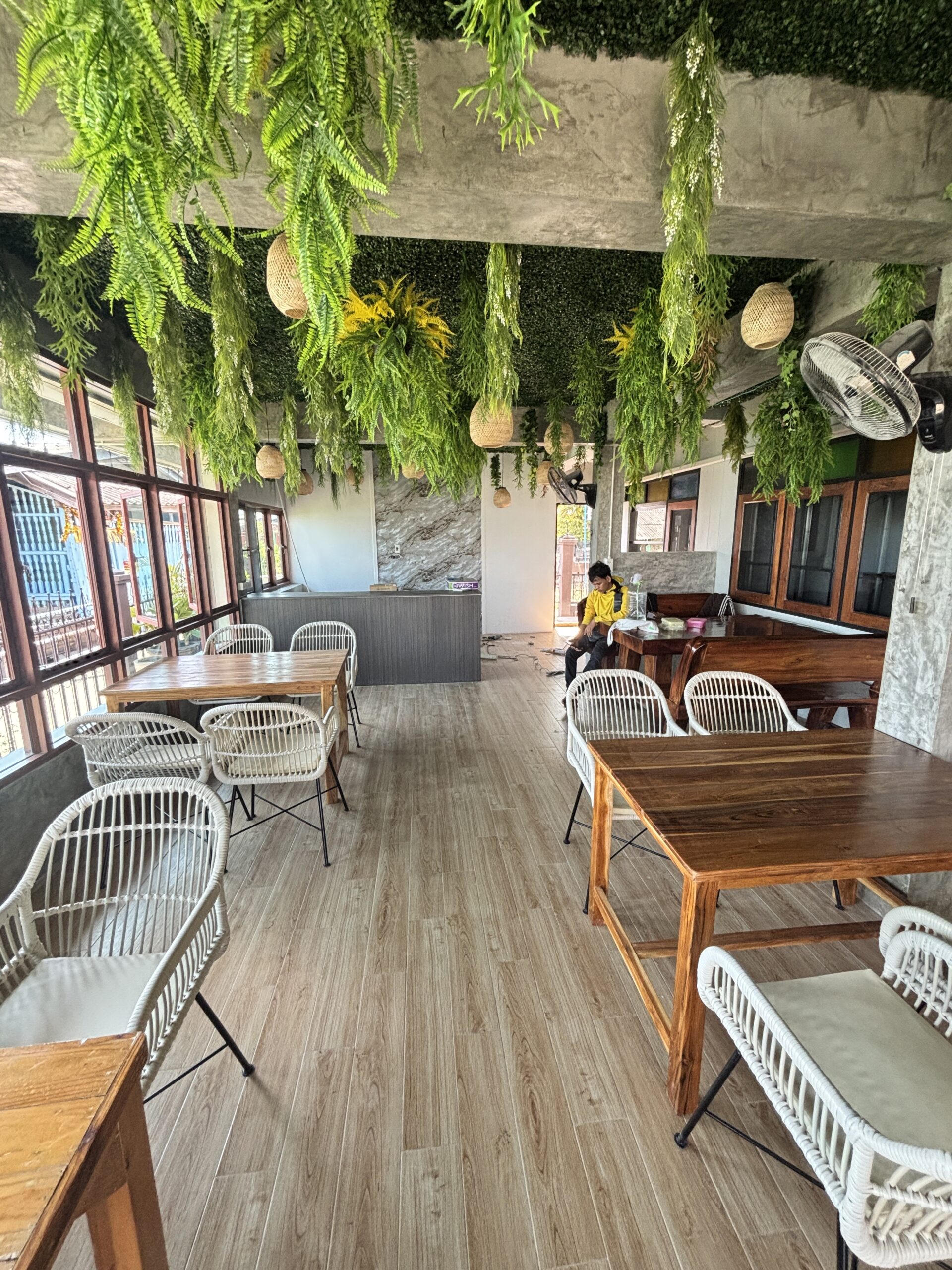 บรรยากาศภายในร้านกาแฟตกแต่งด้วยเฟิร์นเขียวห้อยจากเพดาน โต๊ะไม้และเก้าอี้หวายสีขาว พื้นไม้ลามิเนต