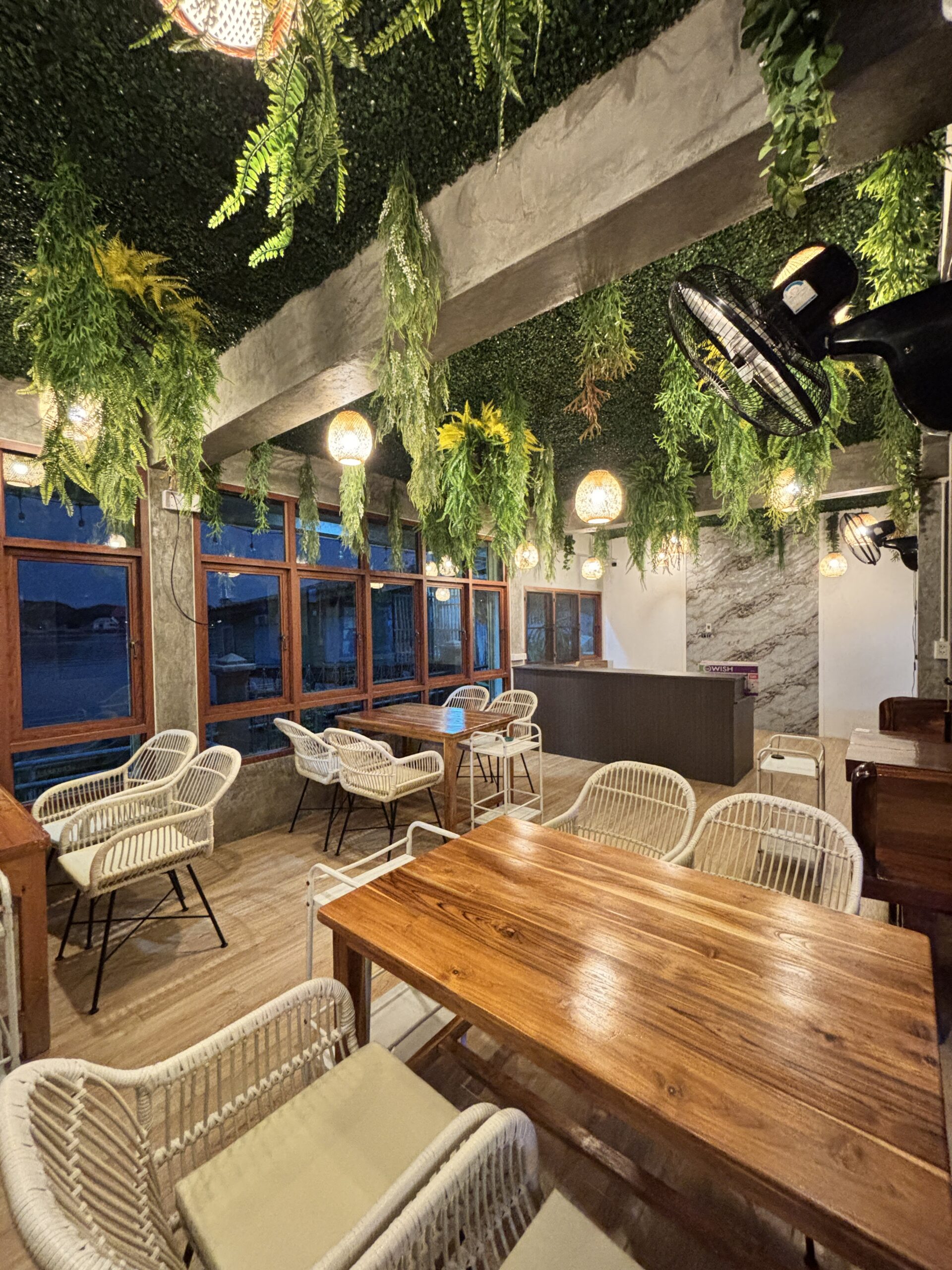 ภายในร้านอาหารตกแต่งสไตล์ทรอปิคอลด้วยต้นเฟิร์นห้อยจากเพดาน โต๊ะไม้และเก้าอี้หวายสีขาว พร้อมไฟแขวนบรรยากาศอบอุ่น