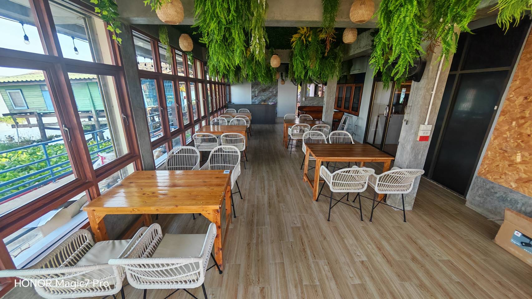 ภายในร้านอาหารตกแต่งสไตล์ทรอปิคอล มีโต๊ะไม้และเก้าอี้หวายสีขาว ประดับด้วยเฟิร์นสีเขียวห้อยจากเพดาน พร้อมหน้าต่างกระจกบานใหญ่รับแสงธรรมชาติ