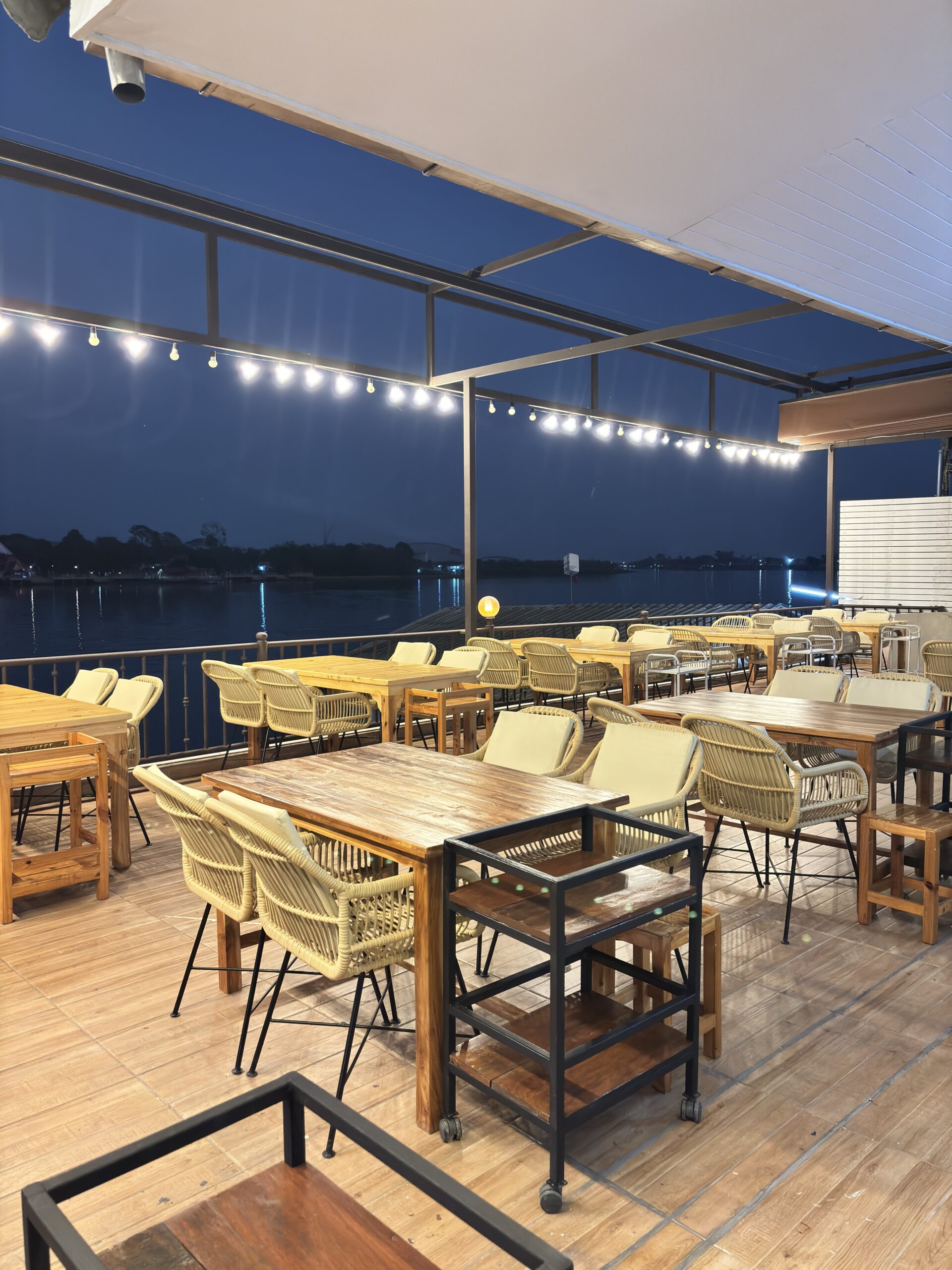 ระเบียงร้านอาหารริมน้ำยามค่ำคืน มีโต๊ะไม้และเก้าอี้หวายพร้อมไฟสายประดับ บรรยากาศสวยงามริมแม่น้ำ