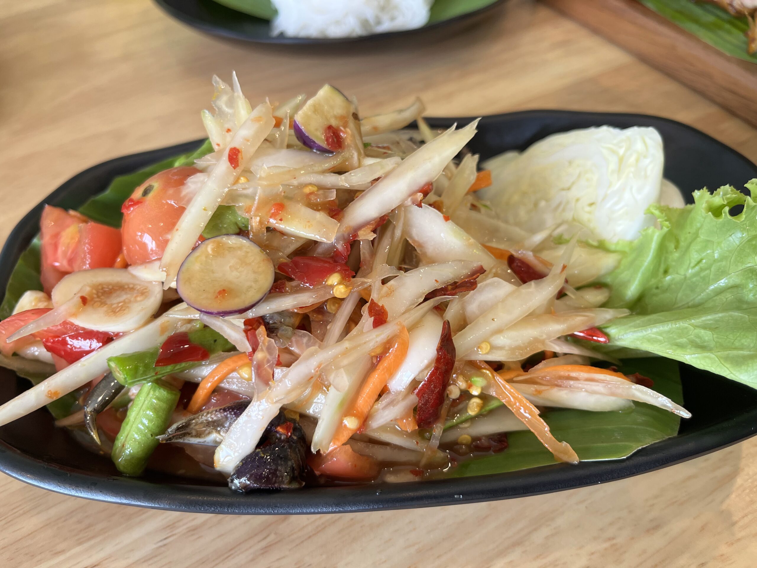 ส้มตำไทยจานใหญ่บนจานดำ มีมะเขือเทศ ถั่วฝักยาว มะเขือเปราะ พริกแห้ง แครอท พร้อมผักสดกะหล่ำปลีและผักกาดแก้ว เสิร์ฟบนใบตอง