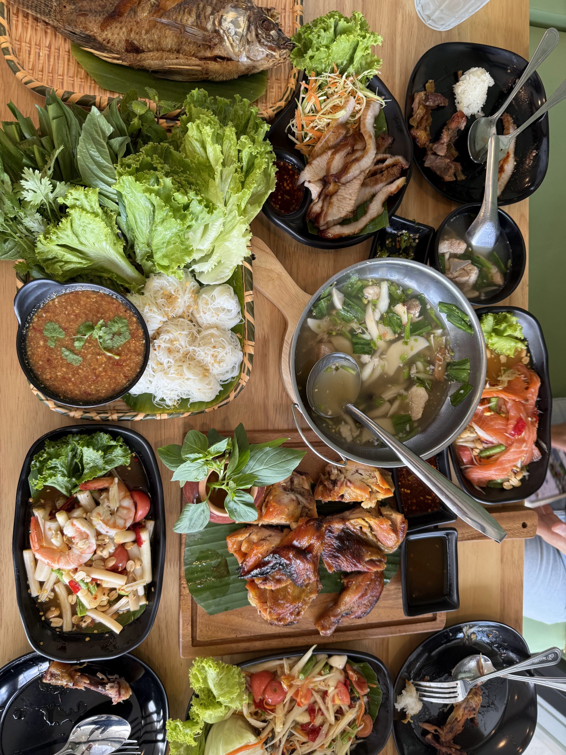 โต๊ะอาหารไทยมองจากด้านบน มีปลาทอด น้ำพริก ขนมจีน ไก่ย่าง ส้มตำ ยำ แกงจืด คอหมูย่าง และผักสดหลากหลายจาน