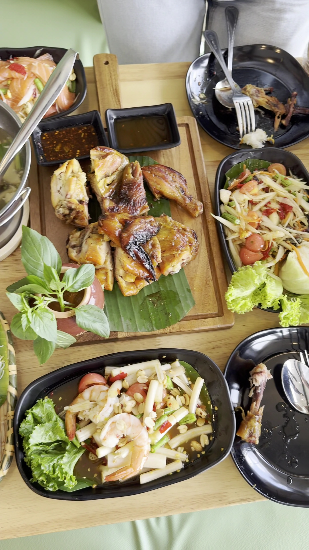 โต๊ะอาหารไทยเต็มไปด้วยไก่ย่างบนใบตอง ส้มตำไทย ยำหัวปลี กุ้ง พร้อมน้ำจิ้มในจานดำ