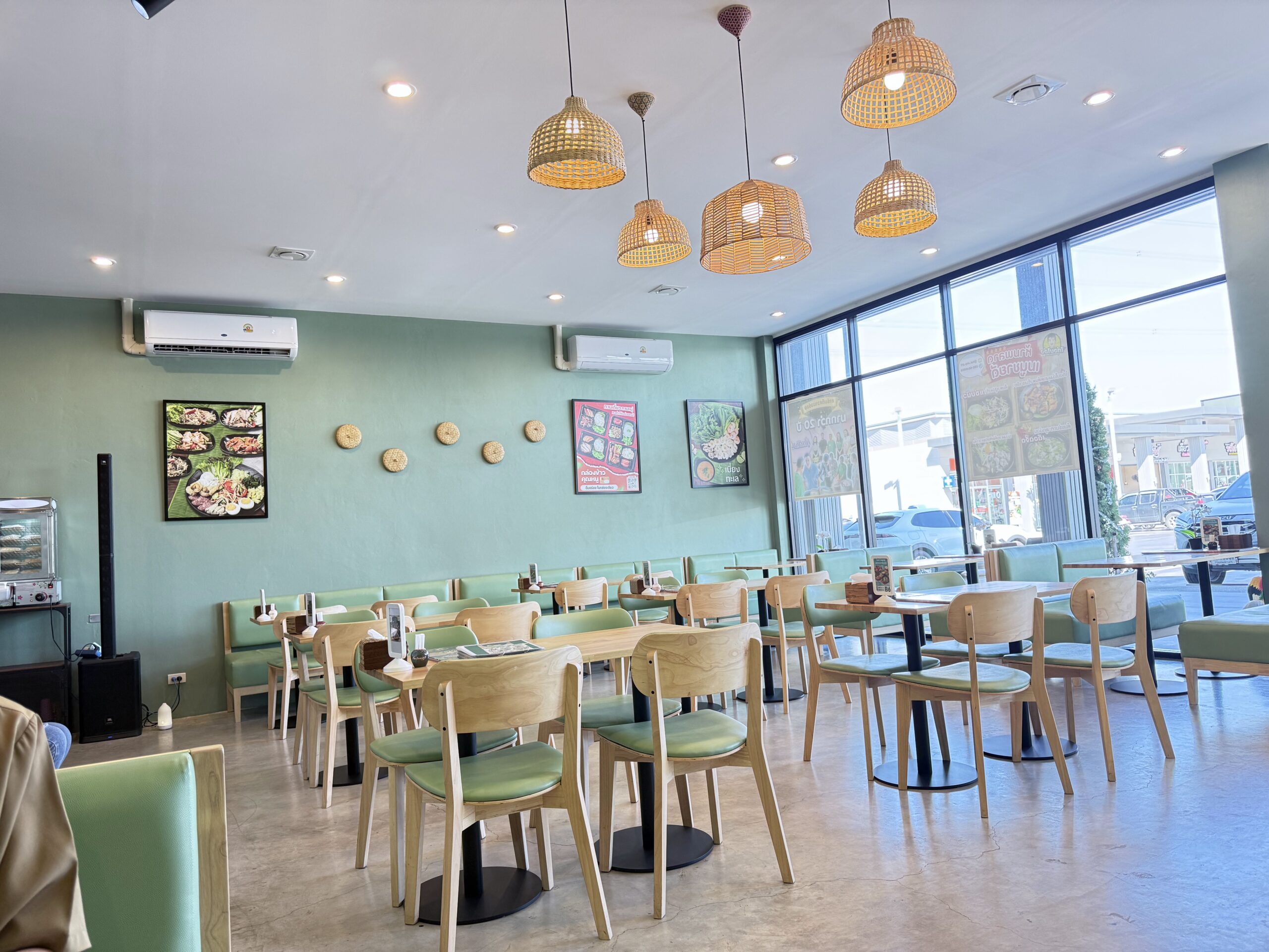 ภายในร้านอาหารไทยตกแต่งโทนสีเขียวมิ้นท์ มีโคมไฟสานไม้ไผ่ เก้าอี้ไม้เบาะสีเขียว โต๊ะไม้ และภาพเมนูอาหารบนผนัง บรรยากาศสะอาดสว่าง