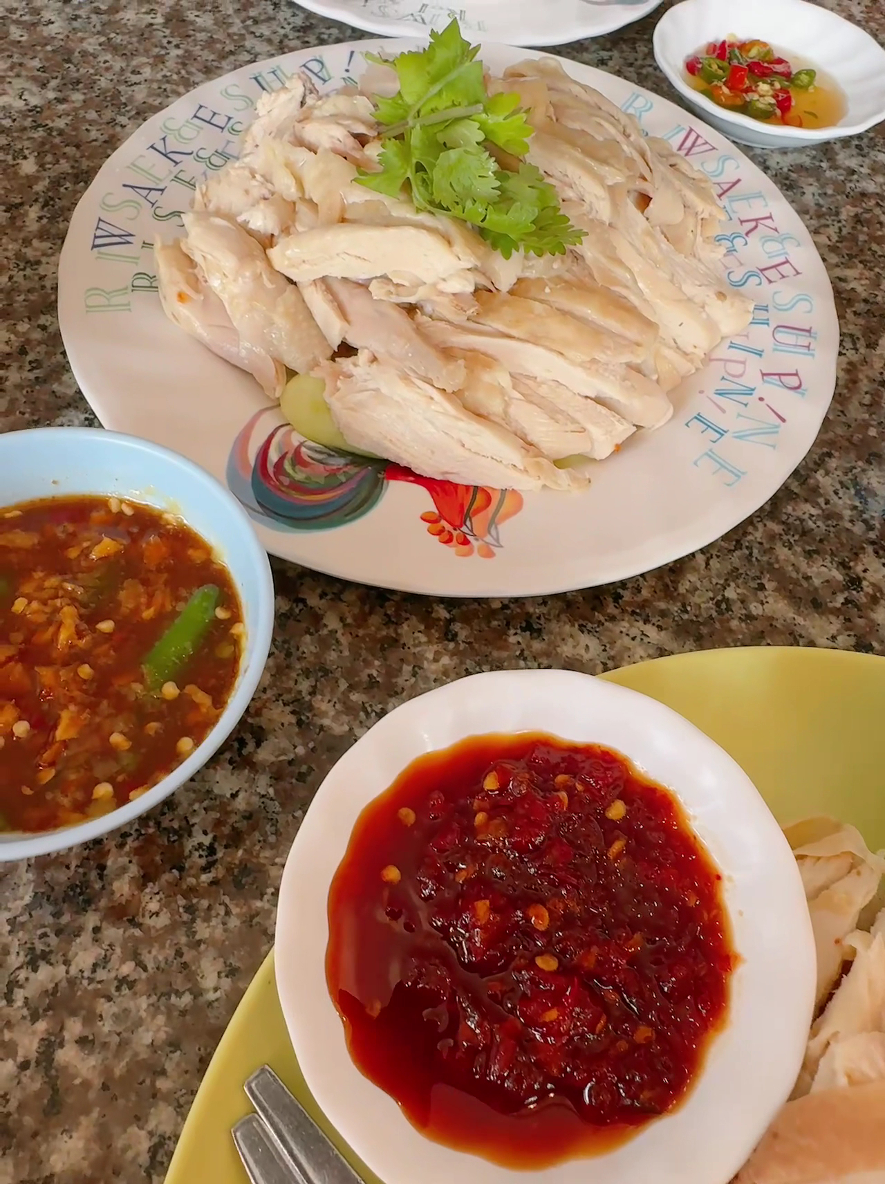 ข้าวมันไก่ต้มหั่นเป็นชิ้นวางบนจานตกแต่งด้วยผักชี พร้อมน้ำจิ้มสามอย่าง ซอสพริกแดง น้ำจิ้มซีอิ๊ว และพริกดองน้ำส้ม