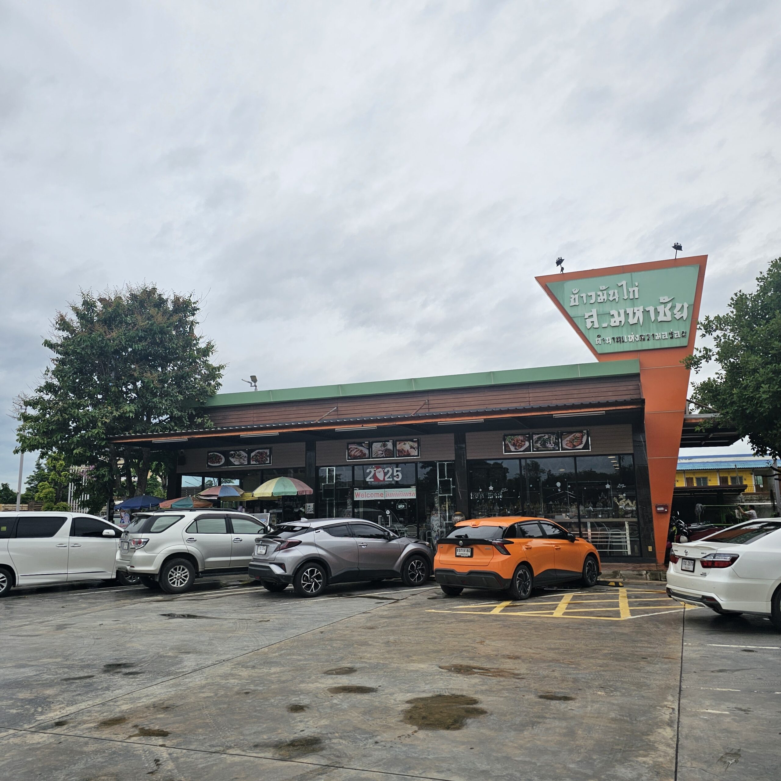 หน้าร้านข้าวมันไก่ ส.มหาชัย พร้อมที่จอดรถกว้างขวาง ป้ายร้านสีส้มและเขียวโดดเด่น มีรถจอดเต็มลานจอด