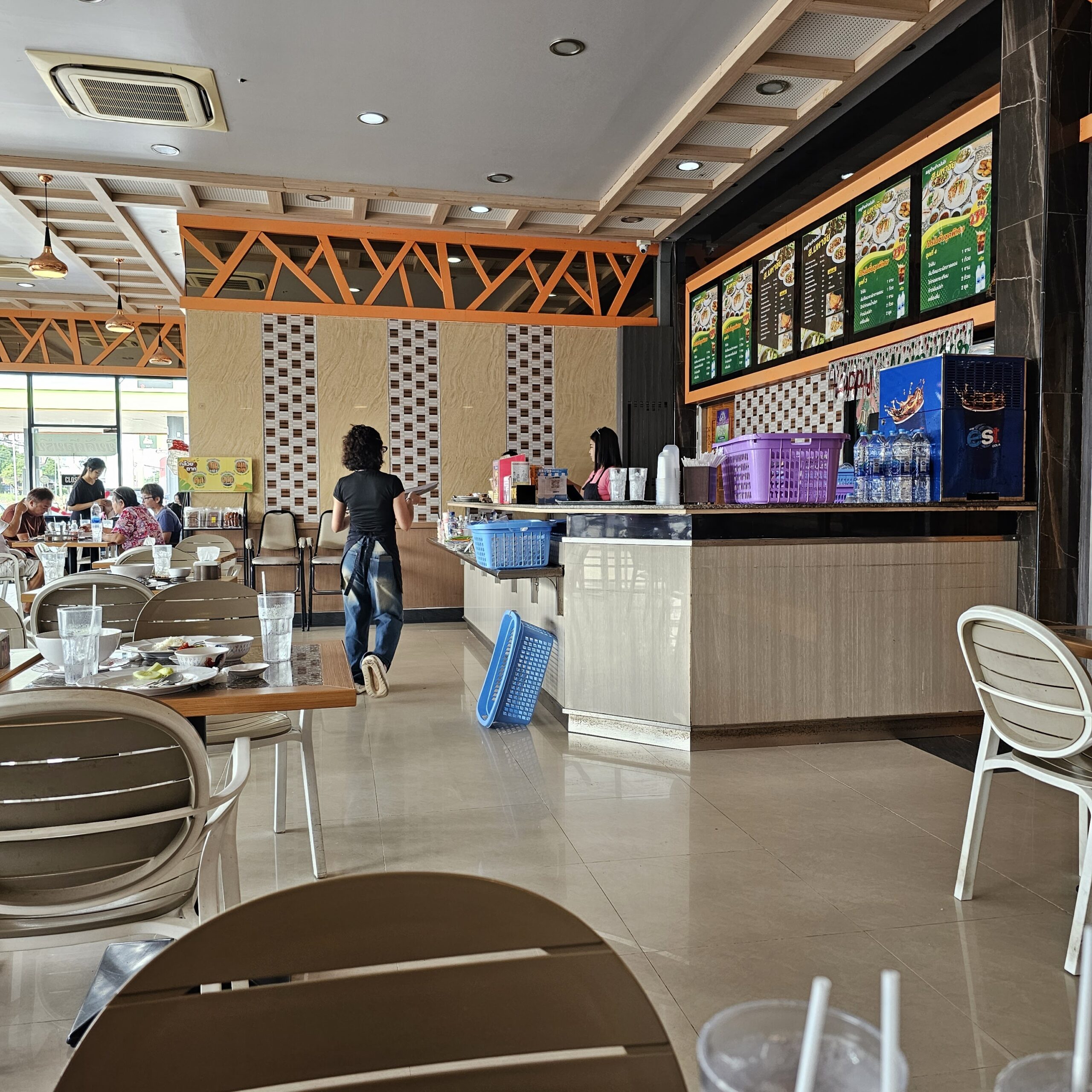 บรรยากาศภายในร้านอาหารไทยพร้อมเคาน์เตอร์สั่งอาหาร เมนูบอร์ดติดผนัง โต๊ะเก้าอี้สำหรับลูกค้า และตะกร้าพลาสติกสีสันสดใส