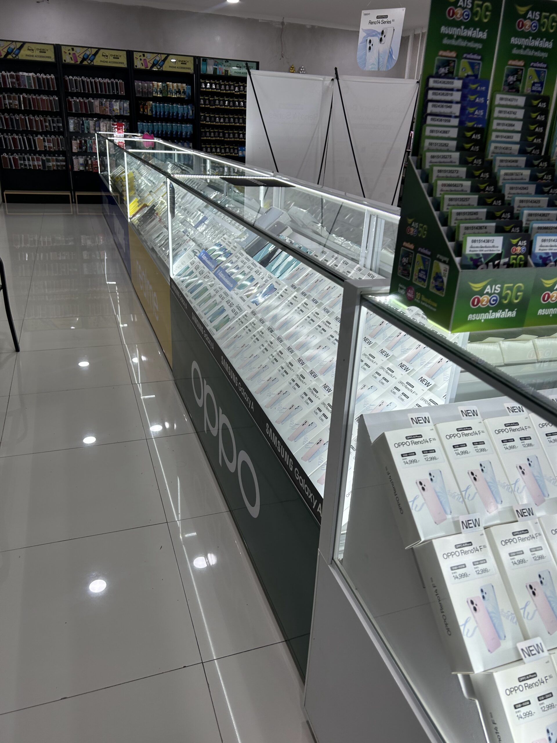 ร้านขายโทรศัพท์มือถือที่มีตู้โชว์กระจกยาวแสดงมือถือหลายยี่ห้อ เช่น OPPO Samsung Galaxy พร้อมป้ายโปรโมชั่น AIS 5G และกล่อง OPPO Reno14 F