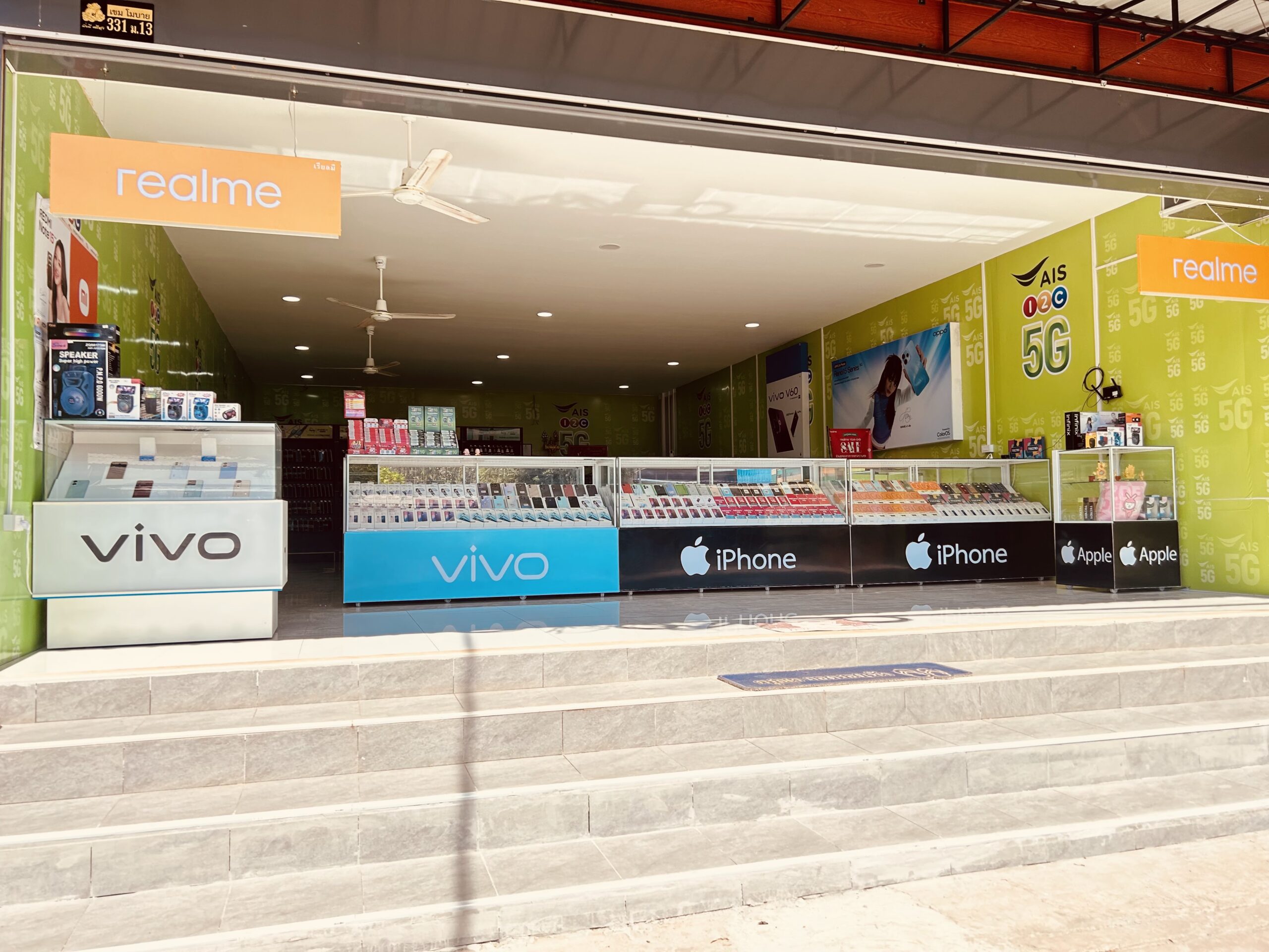 หน้าร้านขายโทรศัพท์มือถือที่มีป้ายแบรนด์ realme, vivo, iPhone และ Apple พร้อมตู้โชว์สินค้าและผนังสีเขียวตกแต่งด้วยลาย 5G