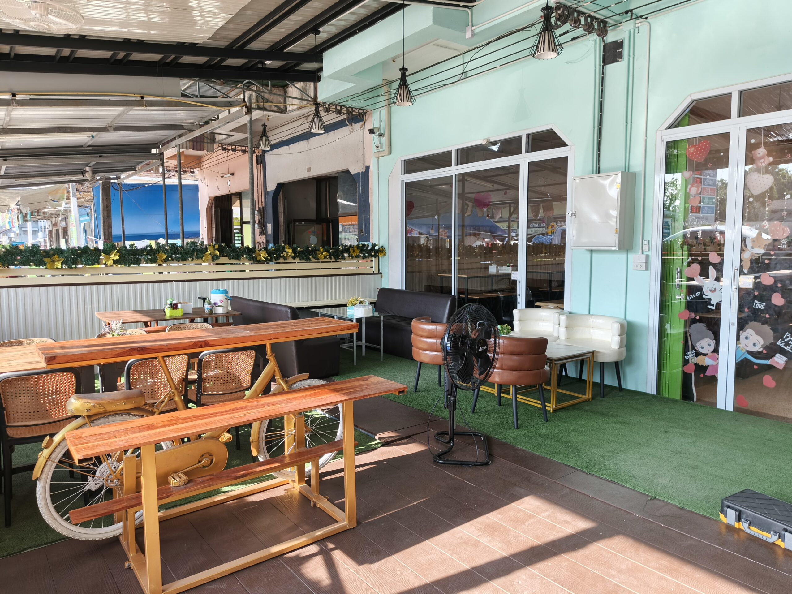 พื้นที่นั่งกลางแจ้งของร้านอาหารตกแต่งด้วยโต๊ะไม้รูปจักรยาน เก้าอี้หวาย โซฟาหนัง พื้นหญ้าเทียมสีเขียว และผนังสีเขียวมิ้นท์