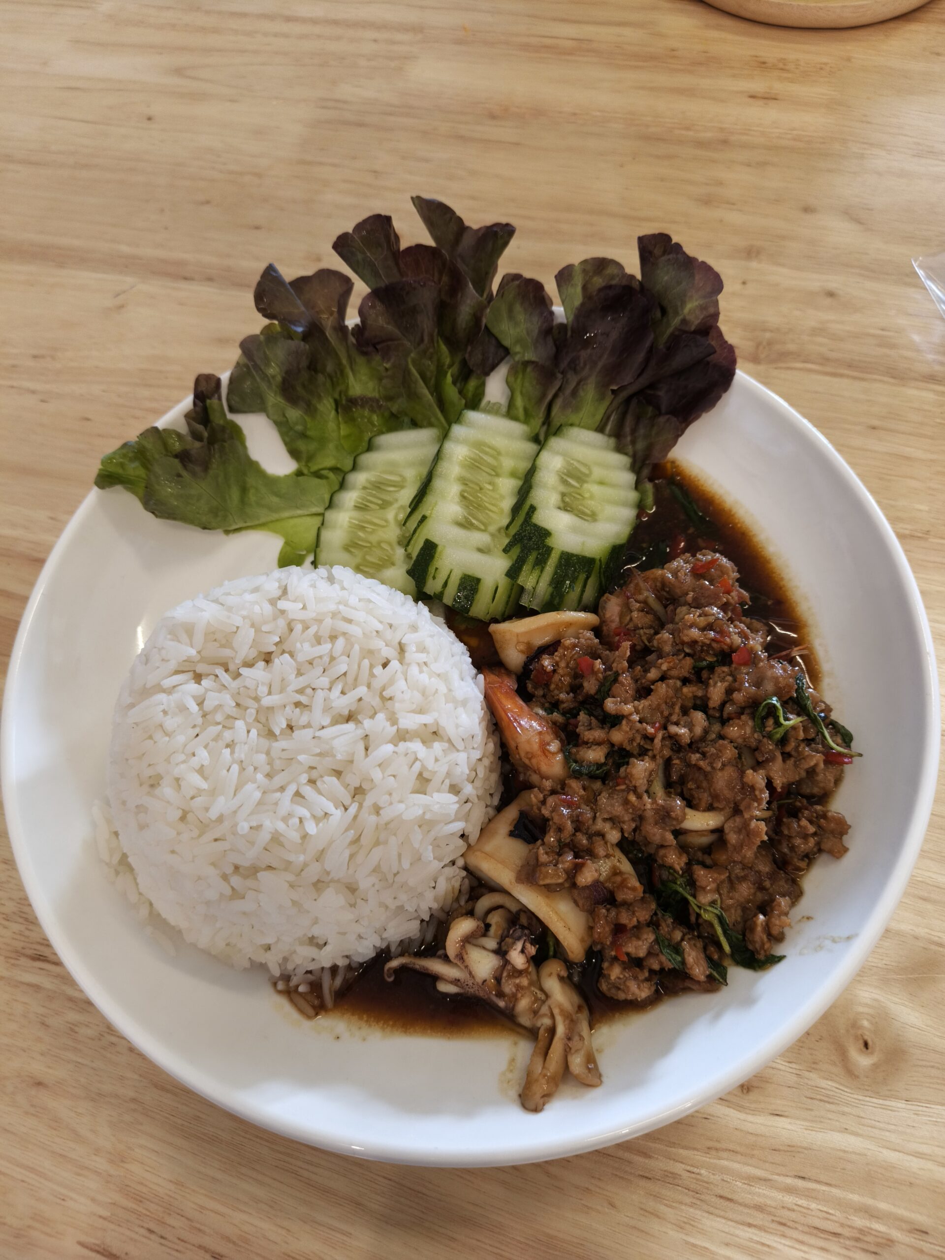 จานข้าวราดกะเพราหมูสับใส่เห็ด พร้อมแตงกวาและผักสลัด เสิร์ฟบนจานขาว