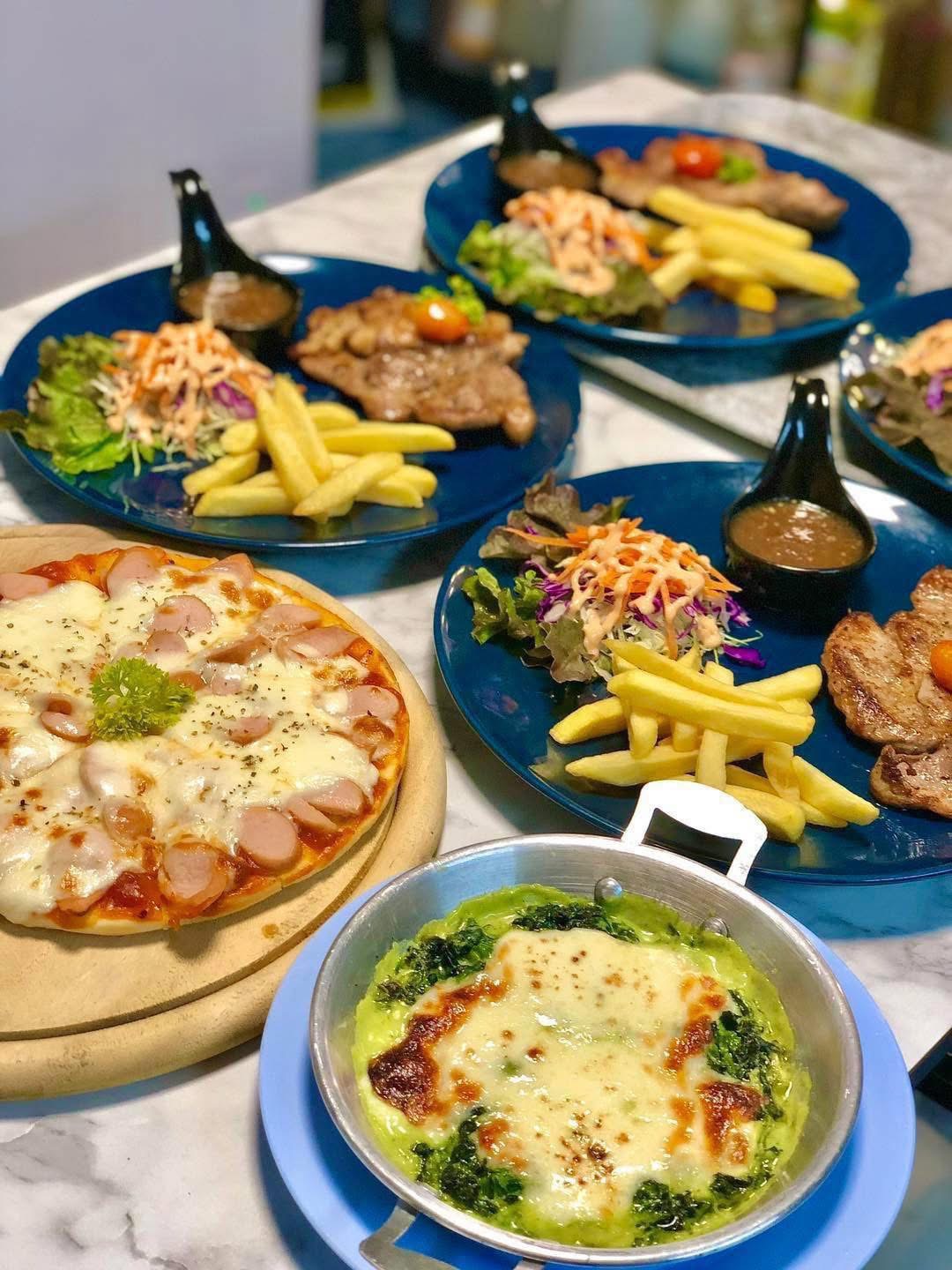 โต๊ะอาหารเต็มไปด้วยจานสีน้ำเงินวางสเต็กหมูพร้อมเฟรนช์ฟรายส์และสลัด พิซซ่าไส้กรอกชีส และสปินาชกราแตงชีสในกระทะ