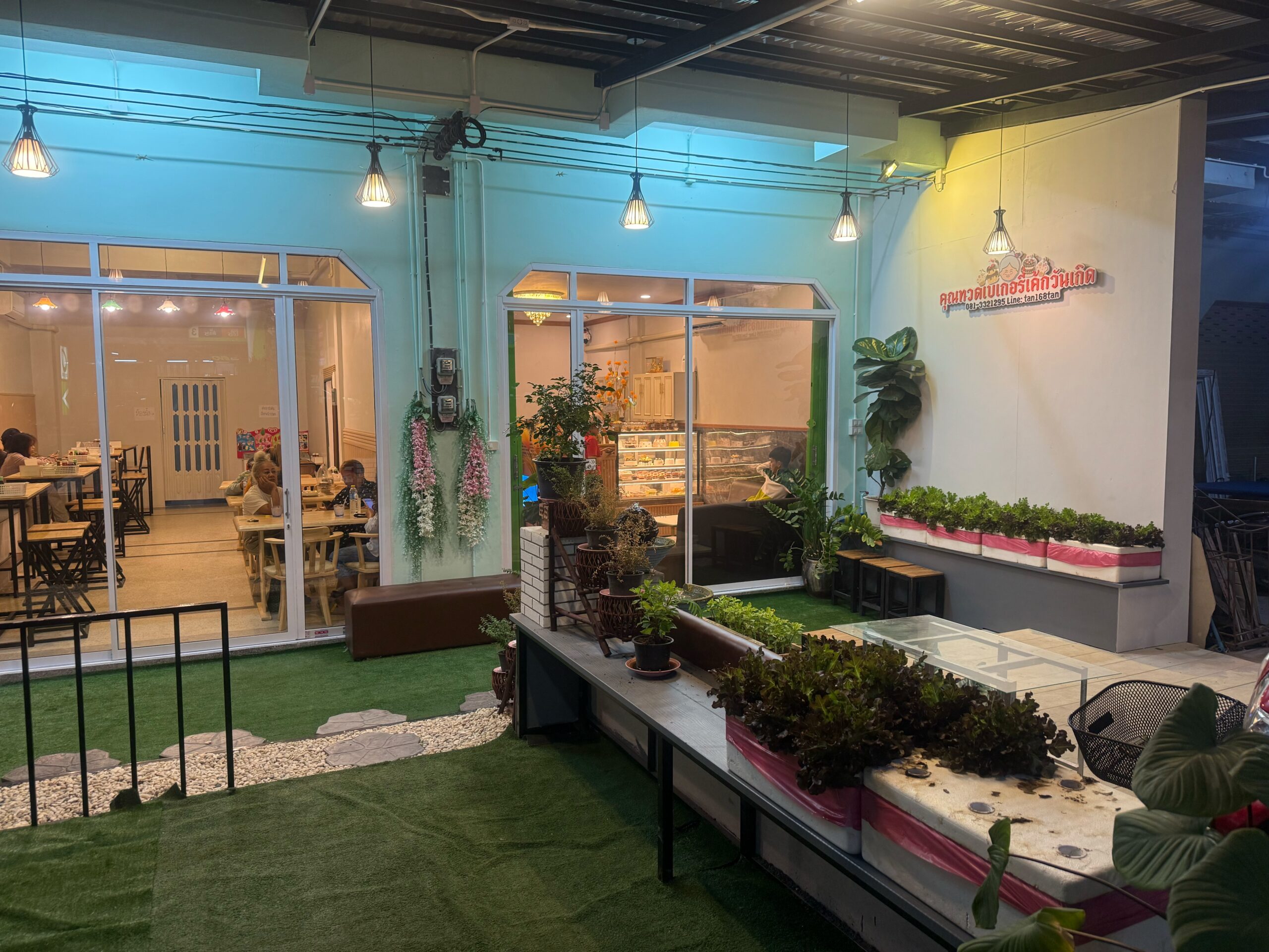 ร้านเบเกอรี่ไทยพร้อมพื้นที่นั่งกลางแจ้งปูหญ้าเทียมสีเขียว มีต้นไม้และไฟตกแต่งสวยงาม มองเห็นตู้โชว์ขนมปังและลูกค้านั่งรับประทานภายในร้าน