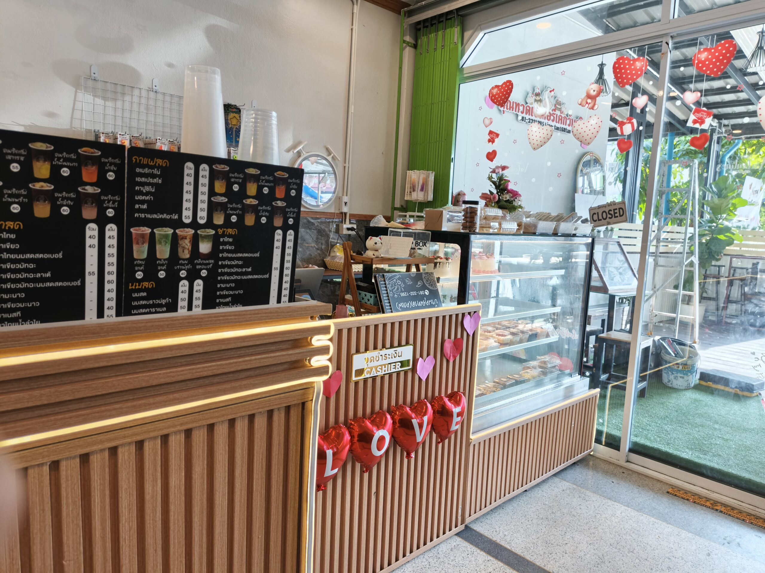 เคาน์เตอร์แคชเชียร์ร้านกาแฟและเบเกอรี่ตกแต่งด้วยลูกโป่งหัวใจสีแดงเขียนคำว่า LOVE มีเมนูกาแฟสดและชาสดติดผนัง พร้อมตู้โชว์เค้กและขนม