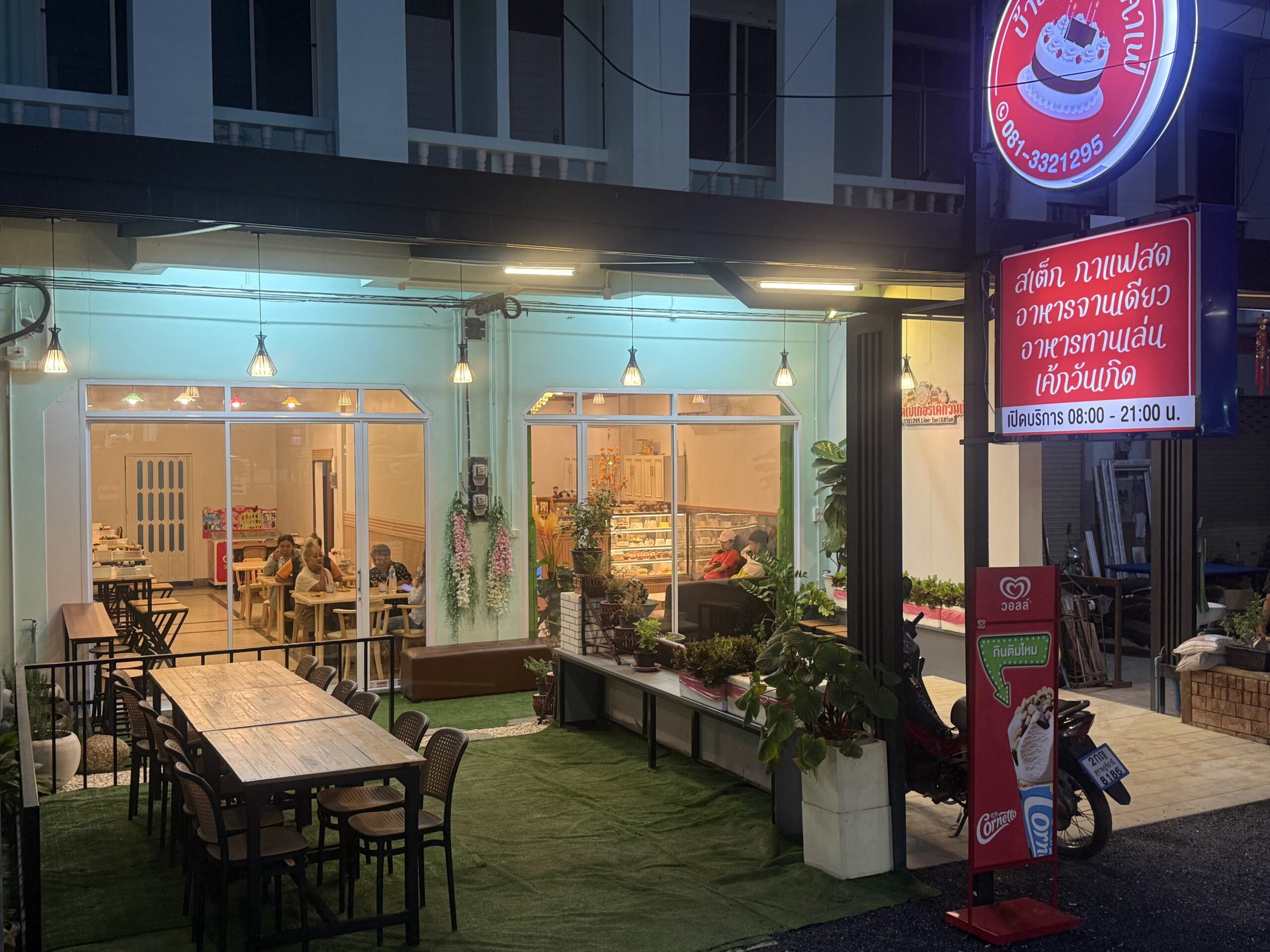 ร้านอาหารไทยยามค่ำคืน มีป้ายสีแดงเขียนว่าสเต็ก กาแฟสด อาหารจานเดียว เค้กวันเกิด เปิดบริการ 08:00-21:00 น. มีโต๊ะนั่งด้านนอกบนหญ้าเทียมสีเขียว และลูกค้านั่งรับประทานอาหารภายในร้าน