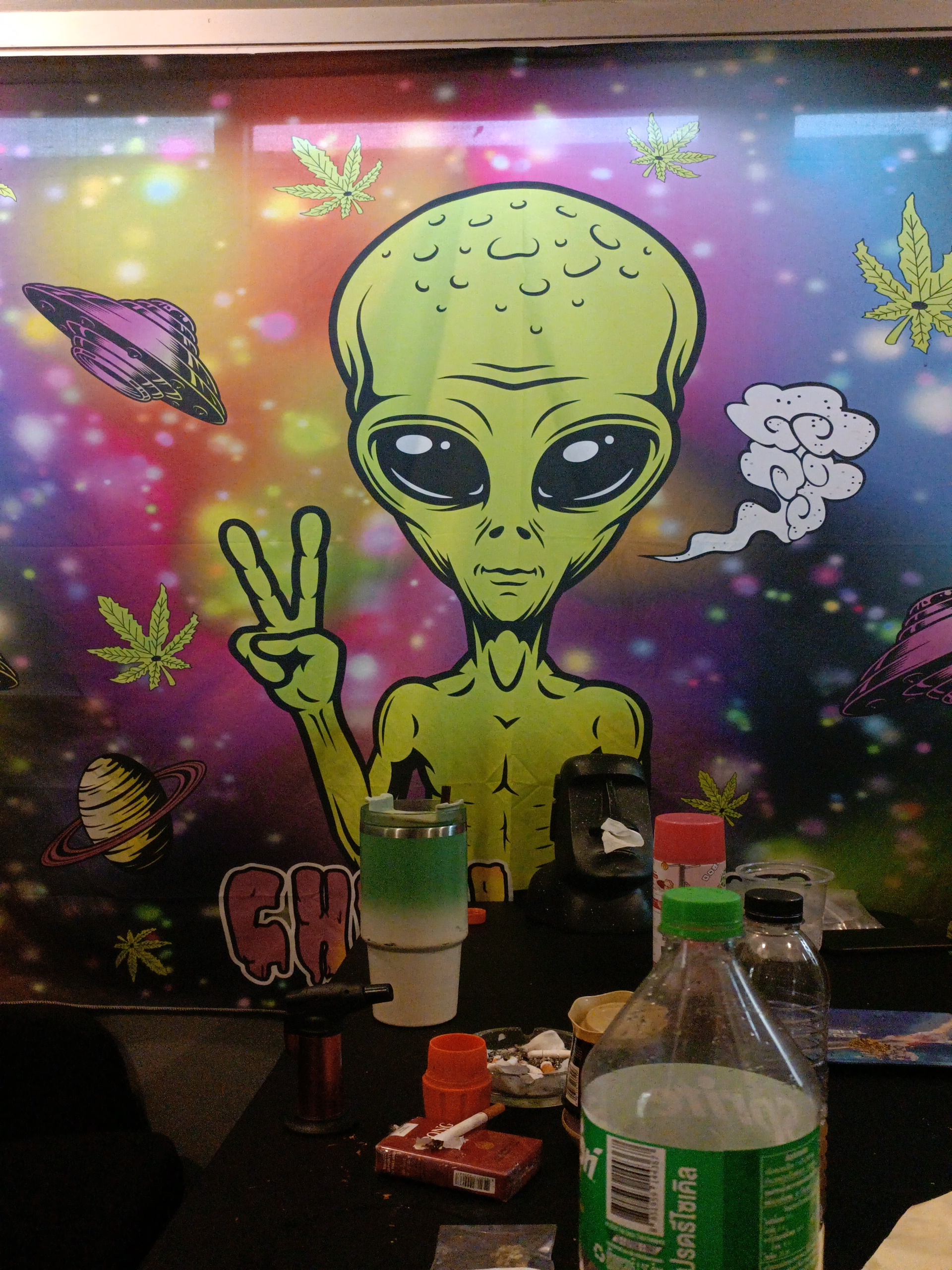 ภาพวาดเอเลี่ยนสีเขียวทำสัญลักษณ์สันติภาพบนผนังร้านที่ตกแต่งธีมอวกาศและใบกัญชา พร้อมขวดน้ำและที่เขี่ยบุหรี่บนโต๊ะ