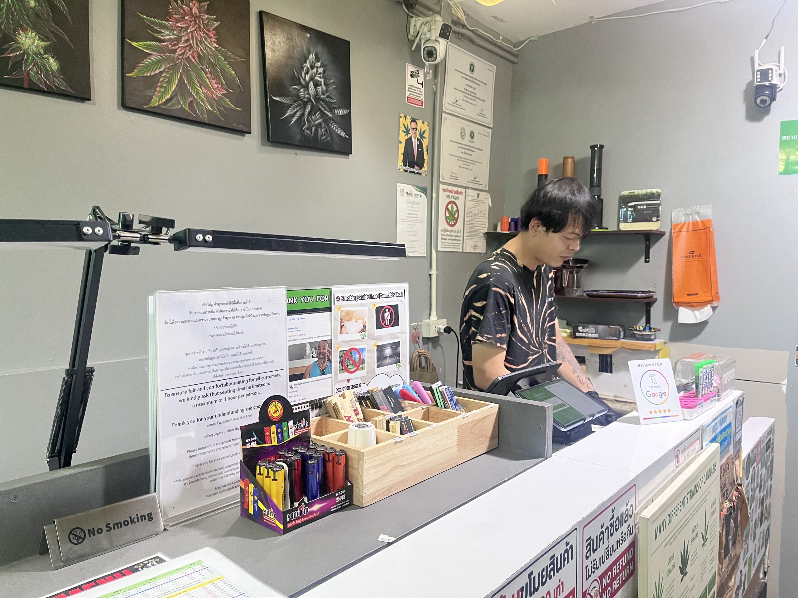 พนักงานชายยืนอยู่หลังเคาน์เตอร์ร้านกัญชาในประเทศไทย มีไฟแช็กหลากสี กระดาษมวน ป้ายห้ามสูบบุหรี่ และภาพวาดใบกัญชาบนผนัง