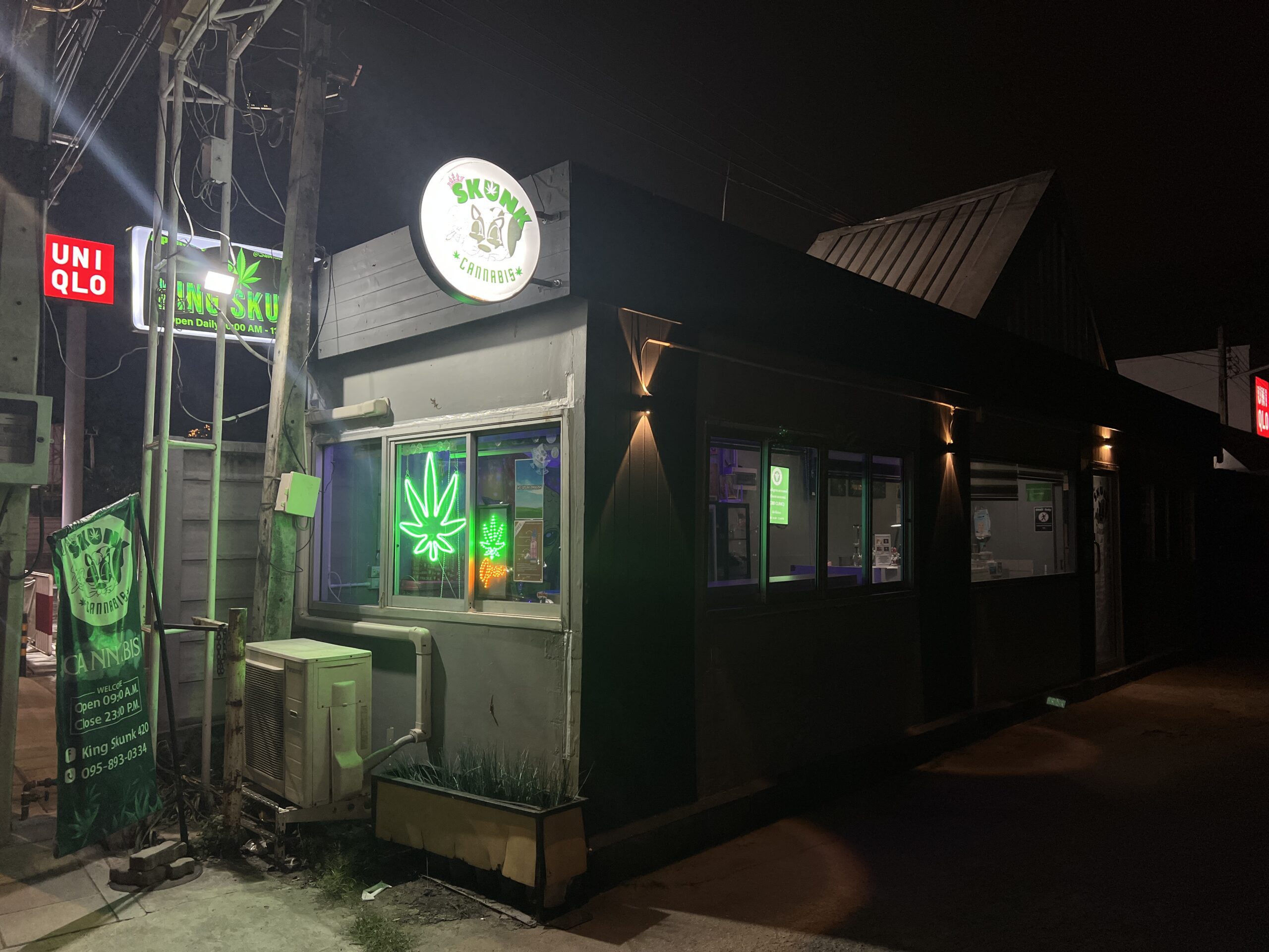 ร้าน King Skunk Cannabis ถ่ายตอนกลางคืน มีป้ายไฟนีออนรูปใบกัญชาสีเขียวและป้ายวงกลมเรืองแสง