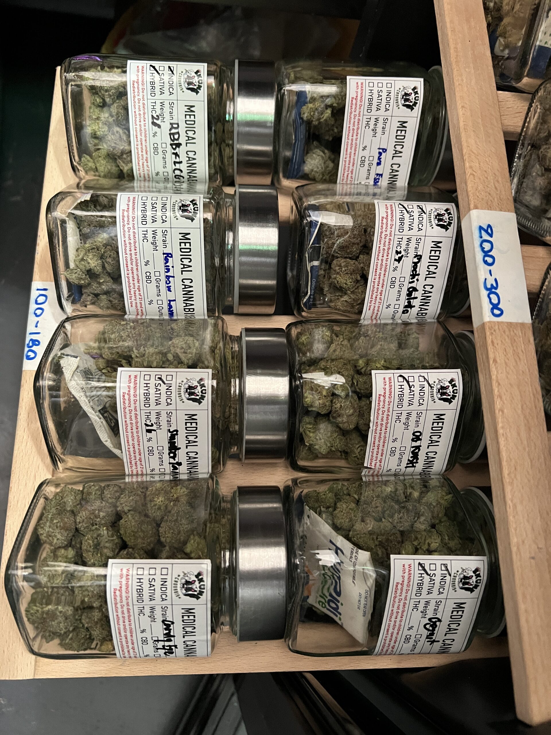 ขวดโหลแก้วบรรจุกัญชาทางการแพทย์หลายสายพันธุ์วางเรียงในชั้นไม้พร้อมฉลากระบุชื่อสายพันธุ์