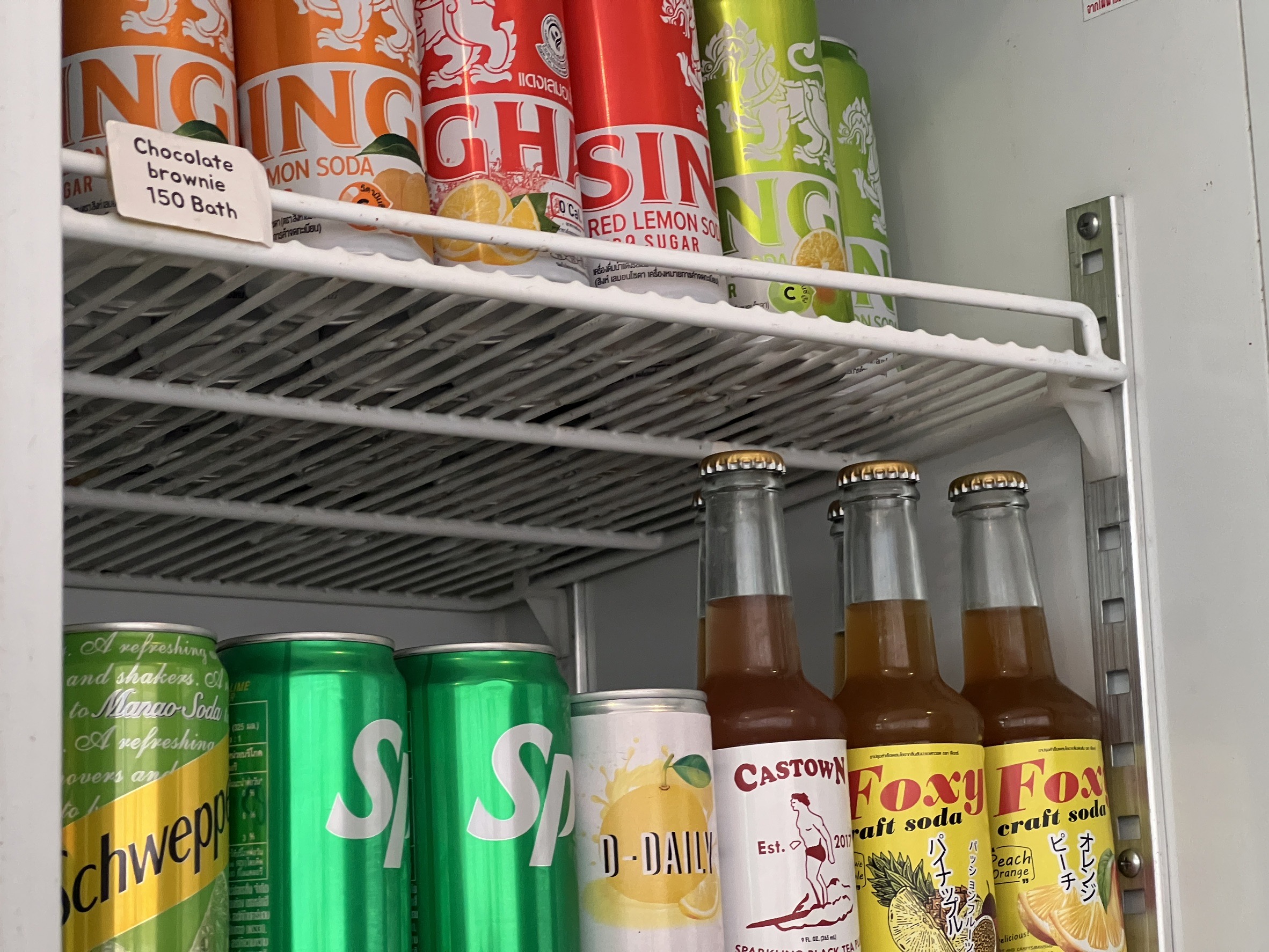 ตู้เย็นแสดงเครื่องดื่มหลากหลายชนิด ได้แก่ น้ำอัดลมสิงห์ เลมอนโซดา ชเวปส์ สไปรท์ Castown และ Foxy craft soda พร้อมป้ายราคาช็อกโกแลตบราวนี่ 150 บาท