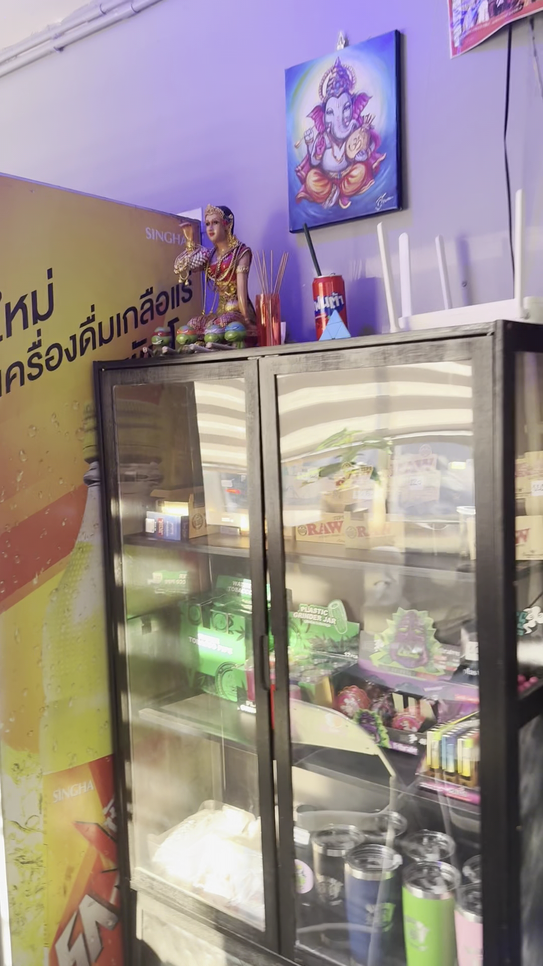 ตู้โชว์สินค้าในร้านค้าไทยพร้อมรูปพระพิฆเนศและตุ๊กตาเทพีไทย มีสินค้ากระดาษมวน RAW และอุปกรณ์ต่างๆ ป้ายเบียร์สิงห์
