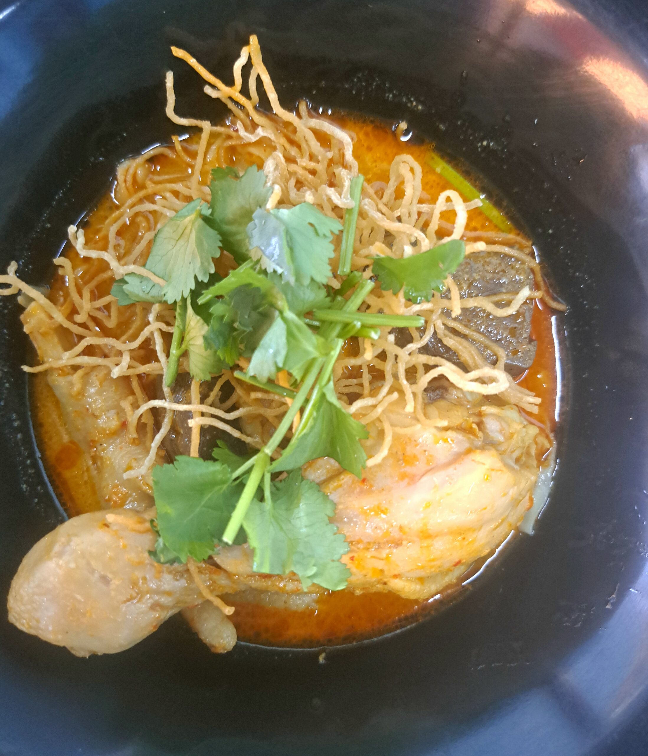 ข้าวซอยไก่ในชามดำ มีน้ำซุปแกงกะหรี่สีส้ม บะหมี่กรอบ น่องไก่ และผักชีโรยหน้า