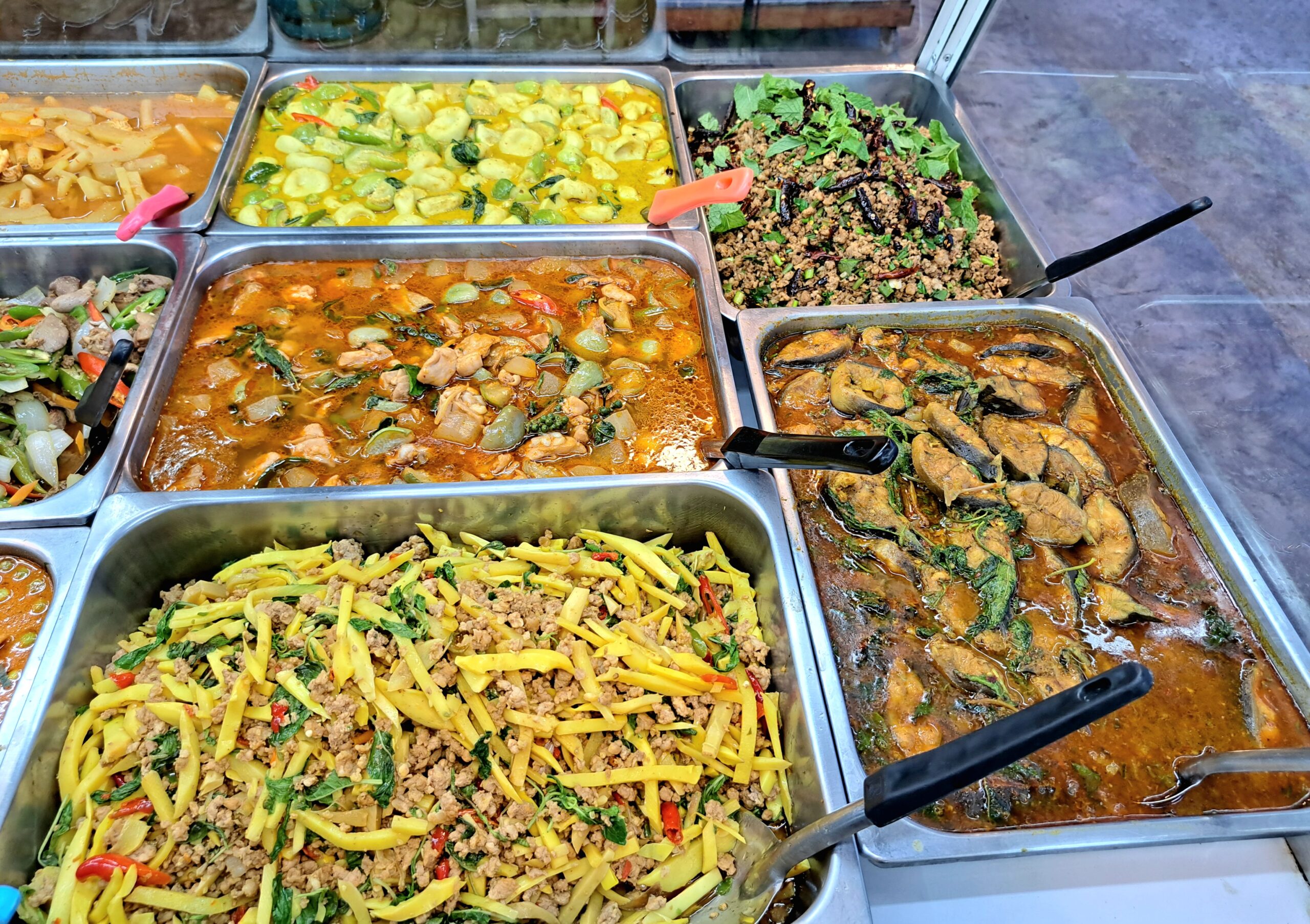 ถาดอาหารตามสั่งหลากหลายเมนู เช่น แกงเผ็ด ลาบ ผัดหน่อไม้ แกงมะเขือ และแกงเขียวหวาน จัดเรียงในถาดสแตนเลสพร้อมเสิร์ฟ