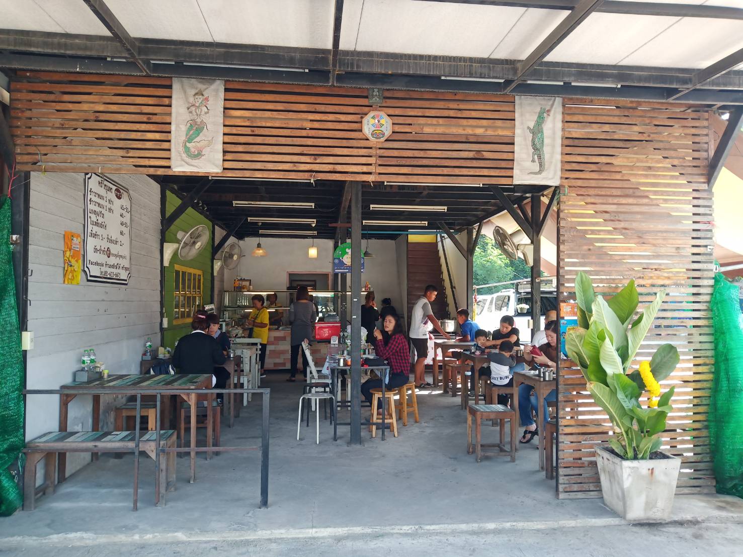 ร้านอาหารไทยแบบเปิดโล่งมีหลังคาไม้ระแนง มีลูกค้านั่งทานอาหารที่โต๊ะไม้ ตกแต่งด้วยป้ายเมนูและผ้าแขวนรูปนางเงือกและจระเข้