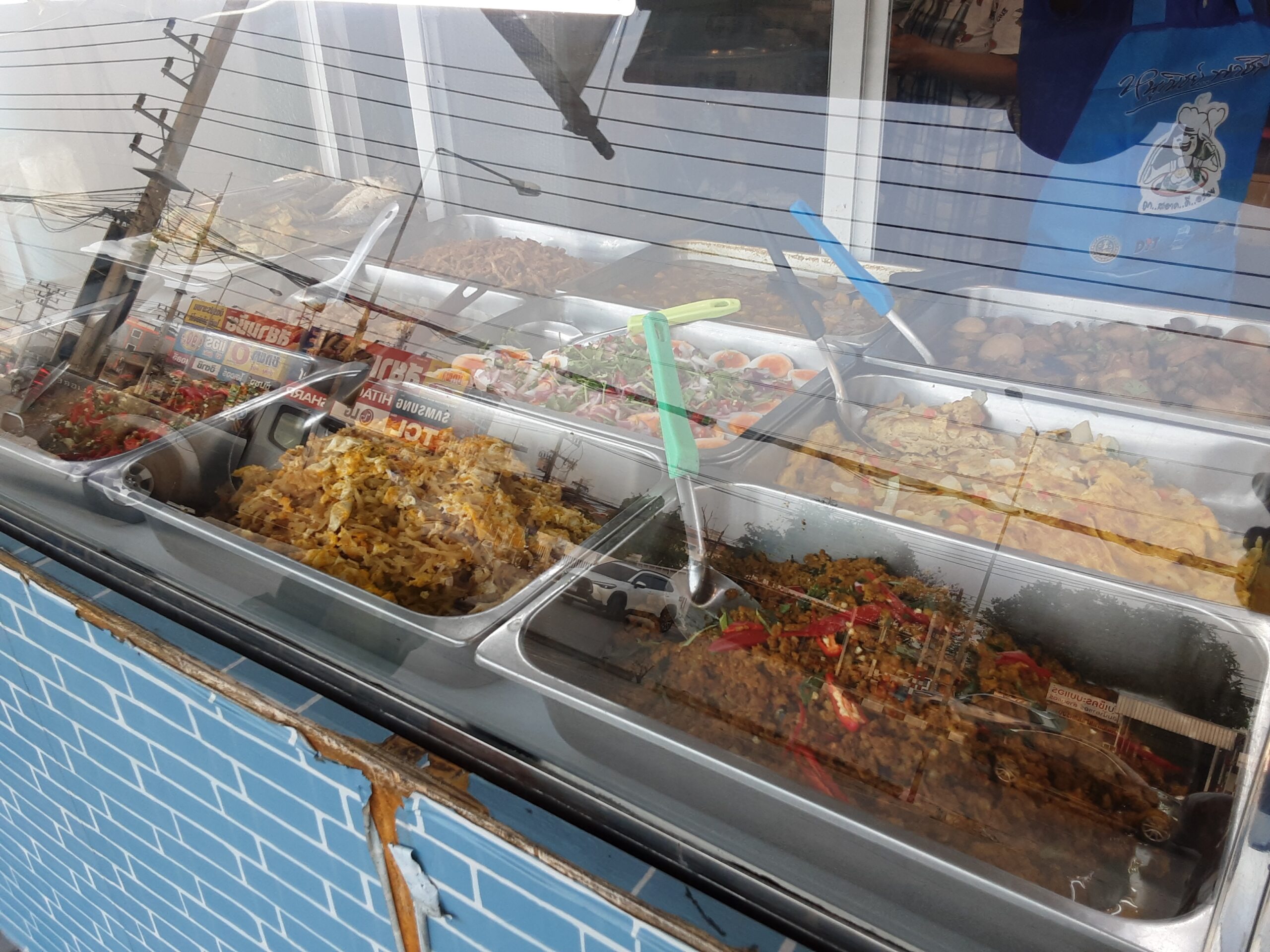 ตู้โชว์อาหารตามสั่งริมถนน มีกับข้าวหลายอย่างในถาดสแตนเลส เช่น ผัดกะเพรา ยำ และแกงต่างๆ พร้อมกระจกกันฝุ่น