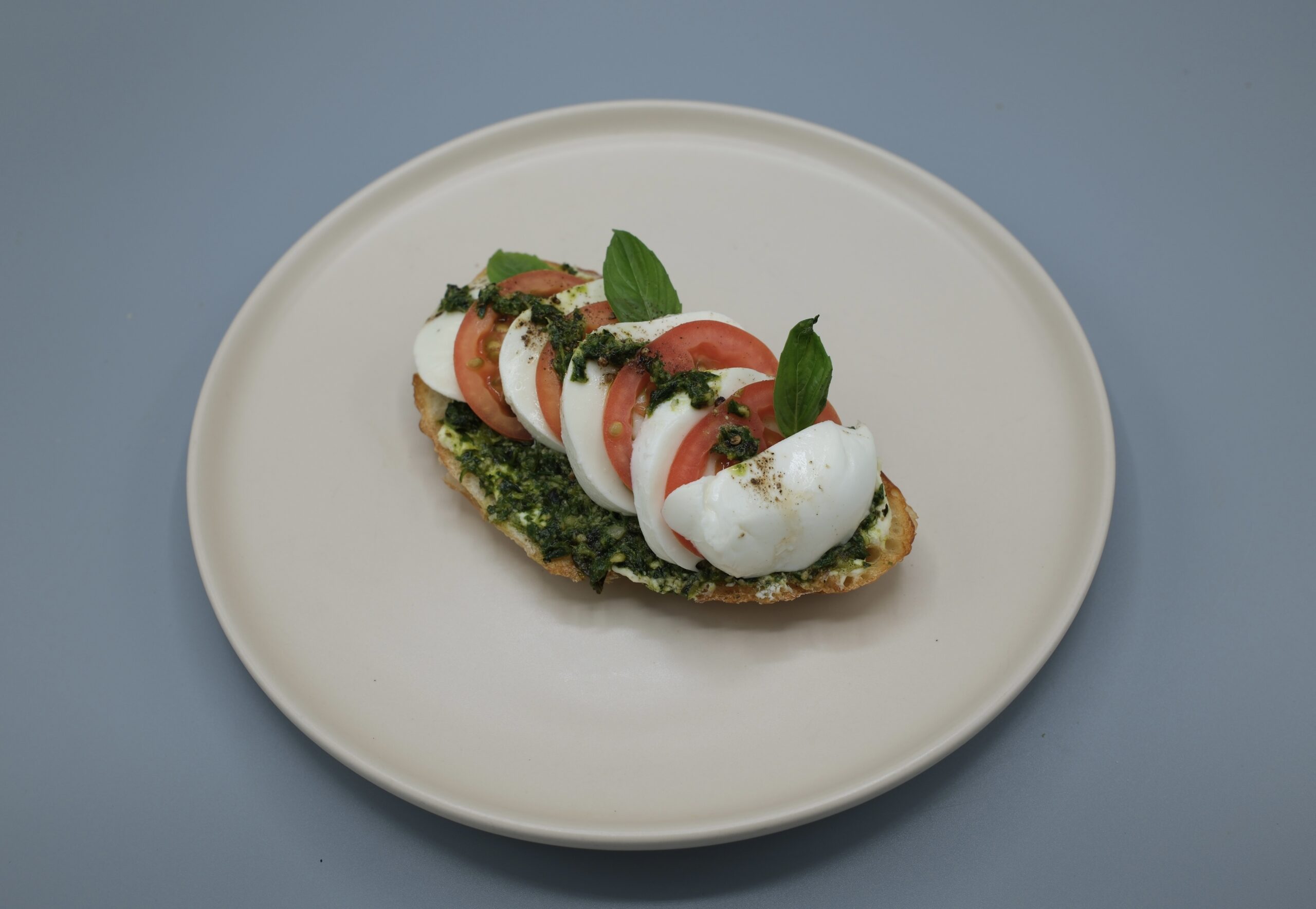 บรูสเก็ตต้าคาปรีเซ่ ขนมปังปิ้งราดซอสเพสโต้ท็อปด้วยมอสซาเรลล่าชีสและมะเขือเทศสดตกแต่งด้วยใบโหระพา บนจานสีขาว