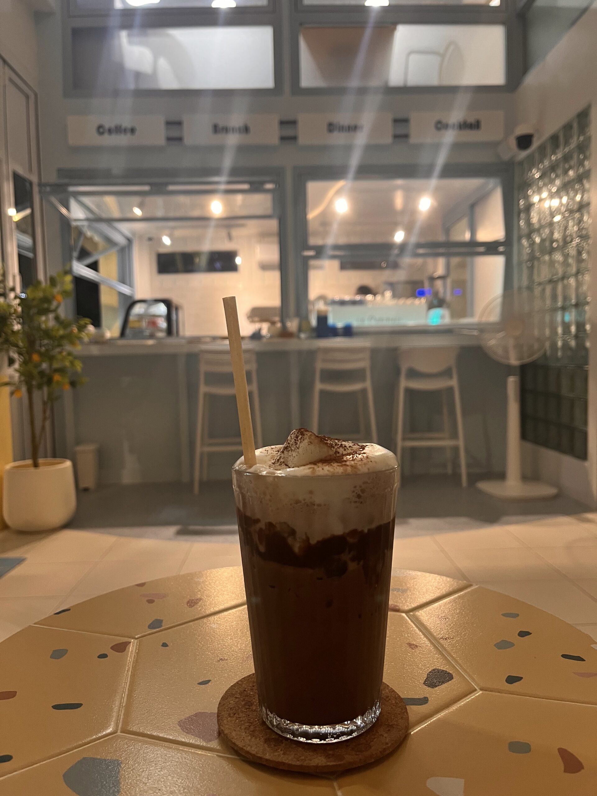 เครื่องดื่มไอซ์มอคค่าท็อปวิปครีมโรยผงโกโก้ วางบนโต๊ะกระเบื้องลายเทอราซโซ่ในร้านกาแฟสไตล์มินิมอลโทนสีขาวเทา