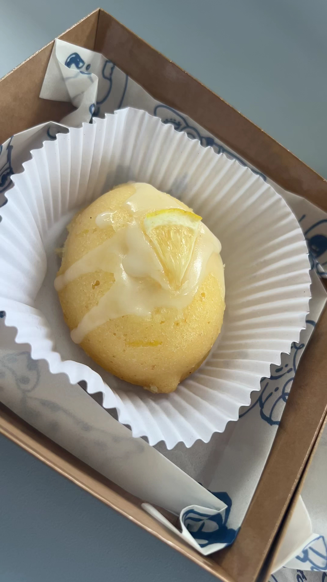 เค้กเลมอนราดน้ำตาลไอซิ่งสีขาวตกแต่งด้วยชิ้นเลมอน วางในถ้วยกระดาษสีขาวในกล่องกระดาษสีน้ำตาล