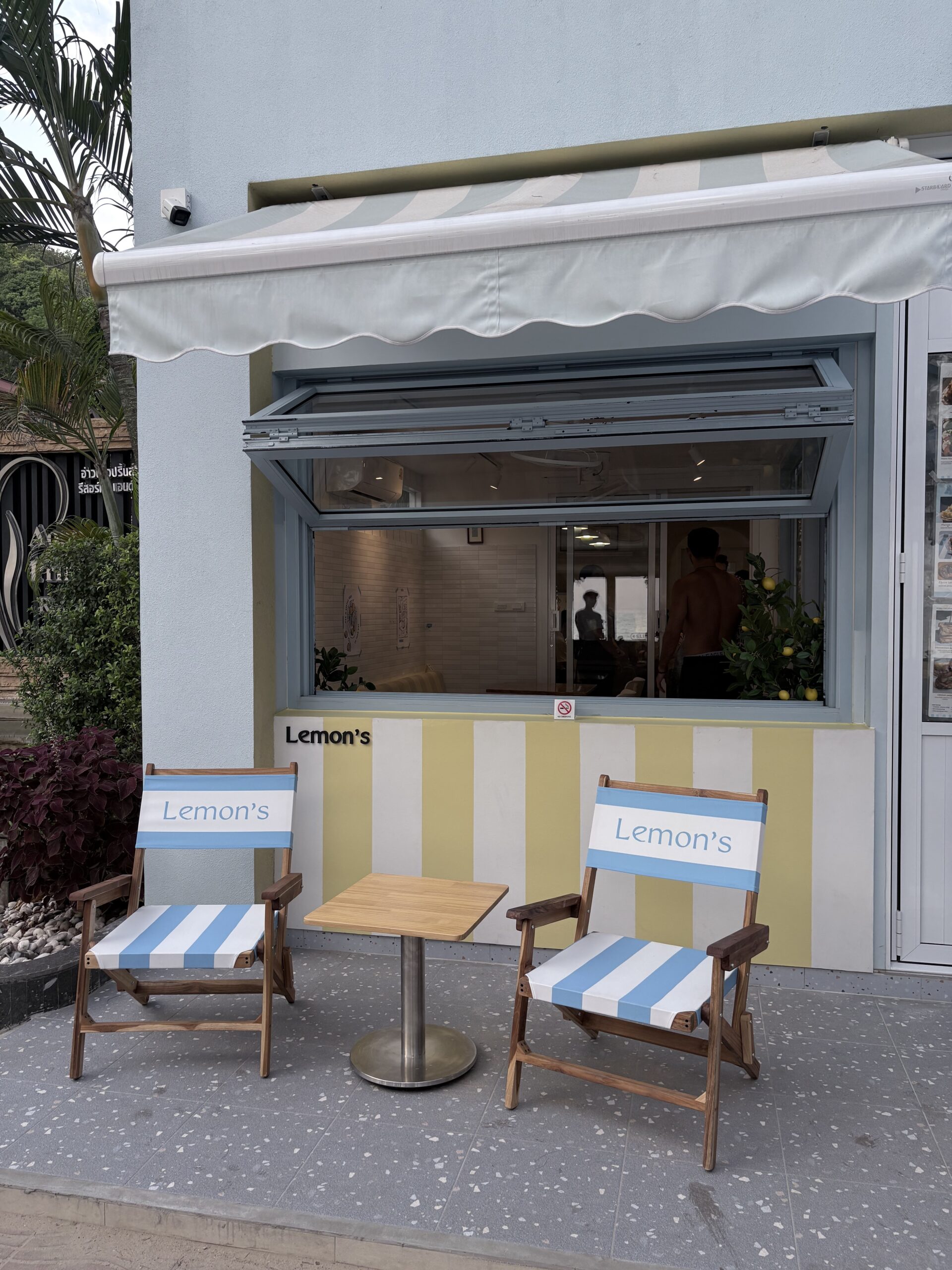 ร้าน Lemon's หน้าร้านสไตล์ชายทะเลพร้อมเก้าอี้ผ้าลายทางสีฟ้าขาว โต๊ะไม้ กันสาดสีขาว และผนังลายทางสีเหลืองครีม