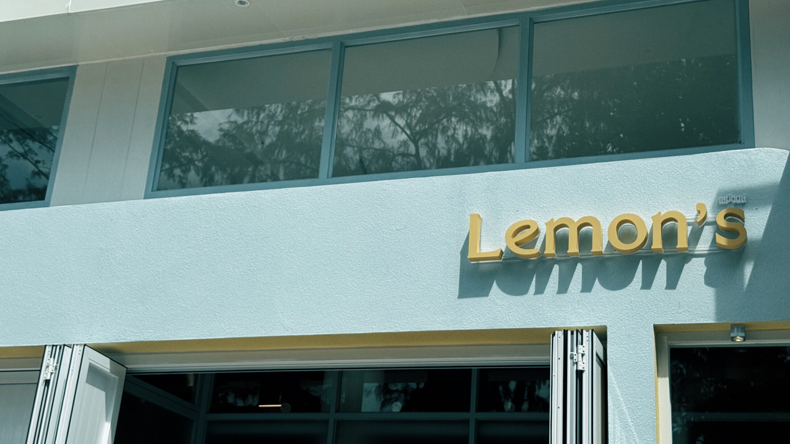 ป้ายชื่อร้าน Lemon's ตัวอักษรสีทองติดบนผนังสีขาวอมฟ้าด้านหน้าร้านอาหาร
