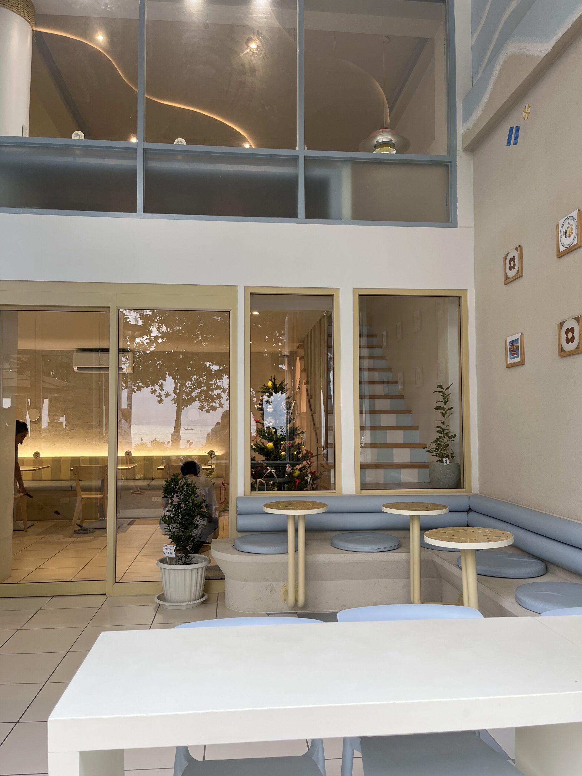 ภายในคาเฟ่สไตล์โมเดิร์นโทนสีขาวและฟ้าพาสเทล มีโซฟานั่งเล่น โต๊ะกลมเล็ก ต้นคริสต์มาส และผนังกระจกสูงสองชั้นพร้อมไฟตกแต่งรูปคลื่น