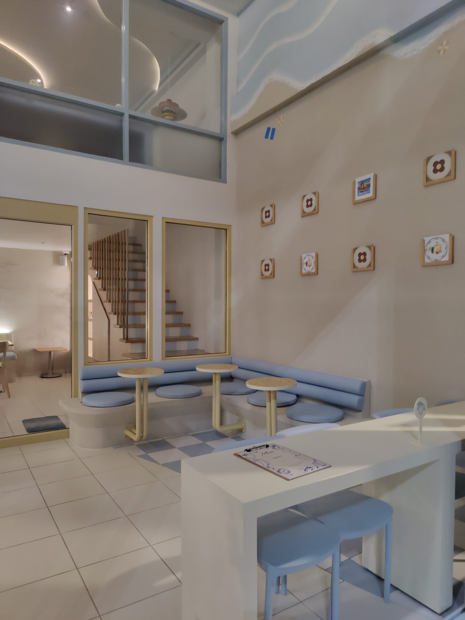 ภายในร้านคาเฟ่โทนสีฟ้าพาสเทลและขาว มีโซฟานั่งยาวสีฟ้า โต๊ะกลมไม้ กรอบรูปตกแต่งผนัง และบันไดไม้มองผ่านกระจก บรรยากาศน่ารักสไตล์กรีก