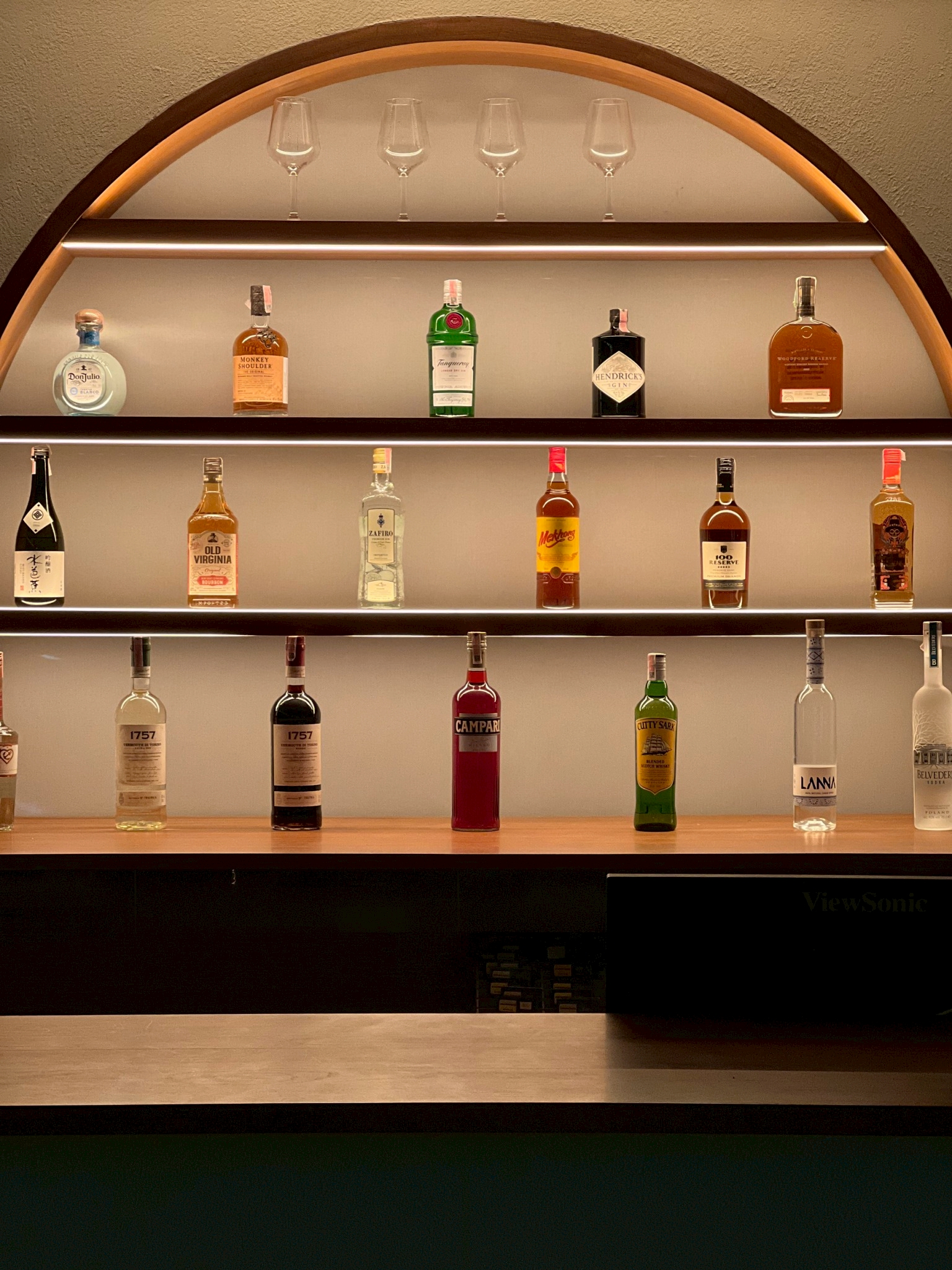 ชั้นวางเครื่องดื่มแอลกอฮอล์หลากชนิดในตู้โค้งมีไฟส่องสว่าง พร้อมแก้วไวน์เรียงอยู่ชั้นบนสุด มีเหล้าหลายยี่ห้อเช่น Don Julio, Monkey Shoulder, Tanqueray, Hendrick's, Campari และ Belvedere