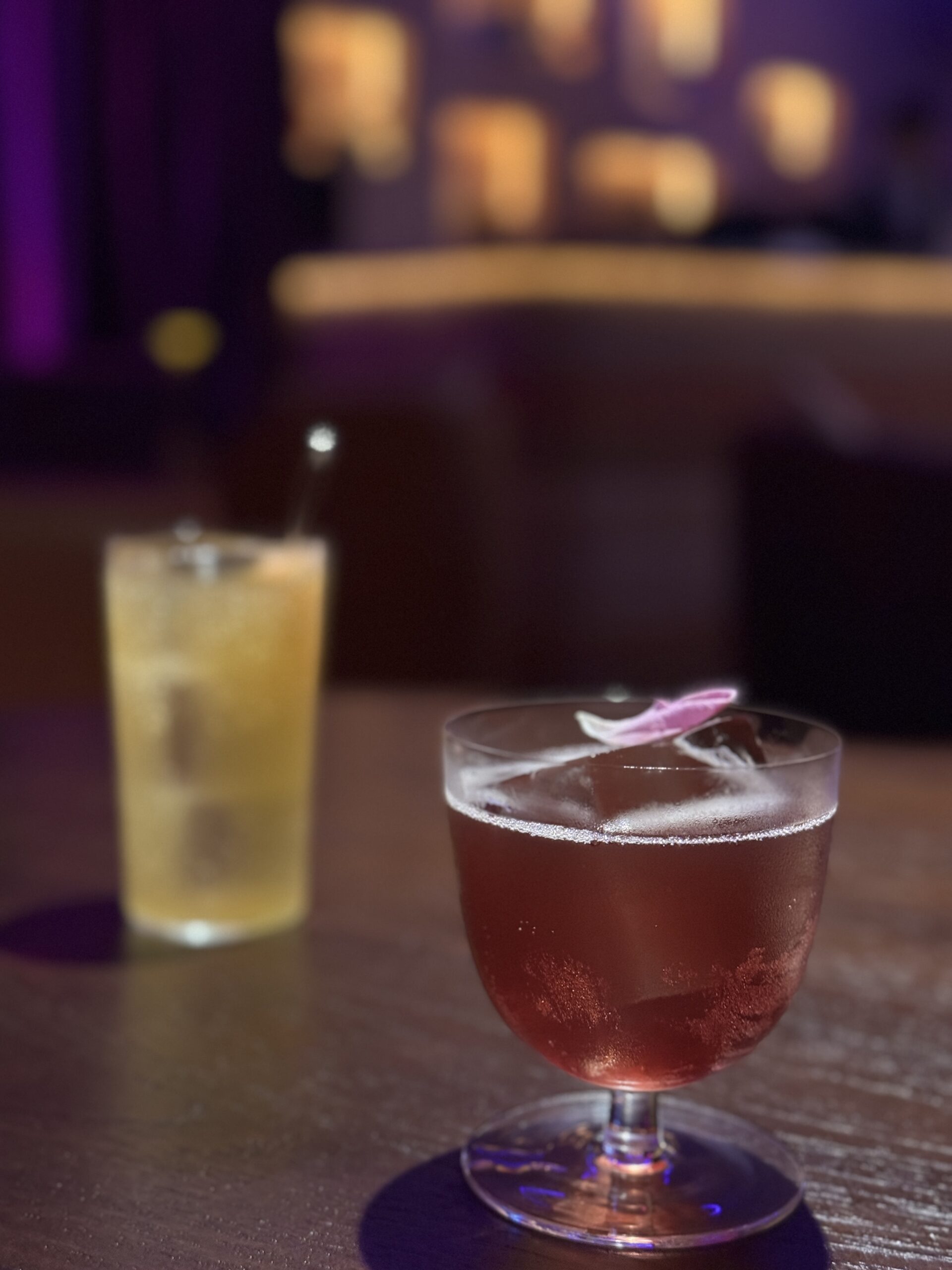 ค็อกเทลสองแก้วบนโต๊ะในบาร์เลาจน์ แก้วหน้าเป็นค็อกเทลสีชมพูแดงตกแต่งด้วยกลีบดอกไม้ แก้วหลังเป็นค็อกเทลสีเหลืองในแก้วทรงสูง บรรยากาศแสงสีม่วงอบอุ่น